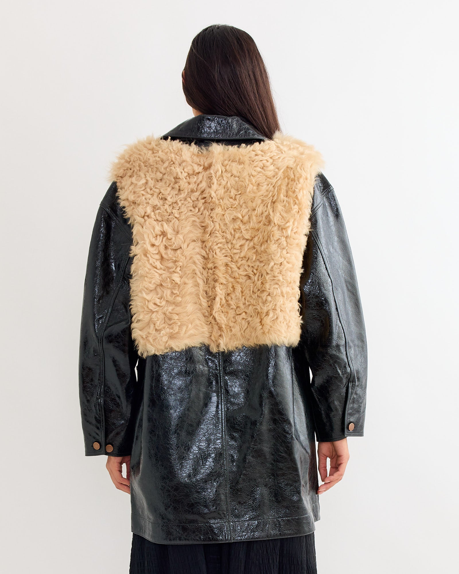 A person with long brown hair, seen from behind, wears a Milena Silvano Chore Coat in Black Mix—a shiny black patent suede jacket with a large tan faux fur panel across the upper back and shoulders.