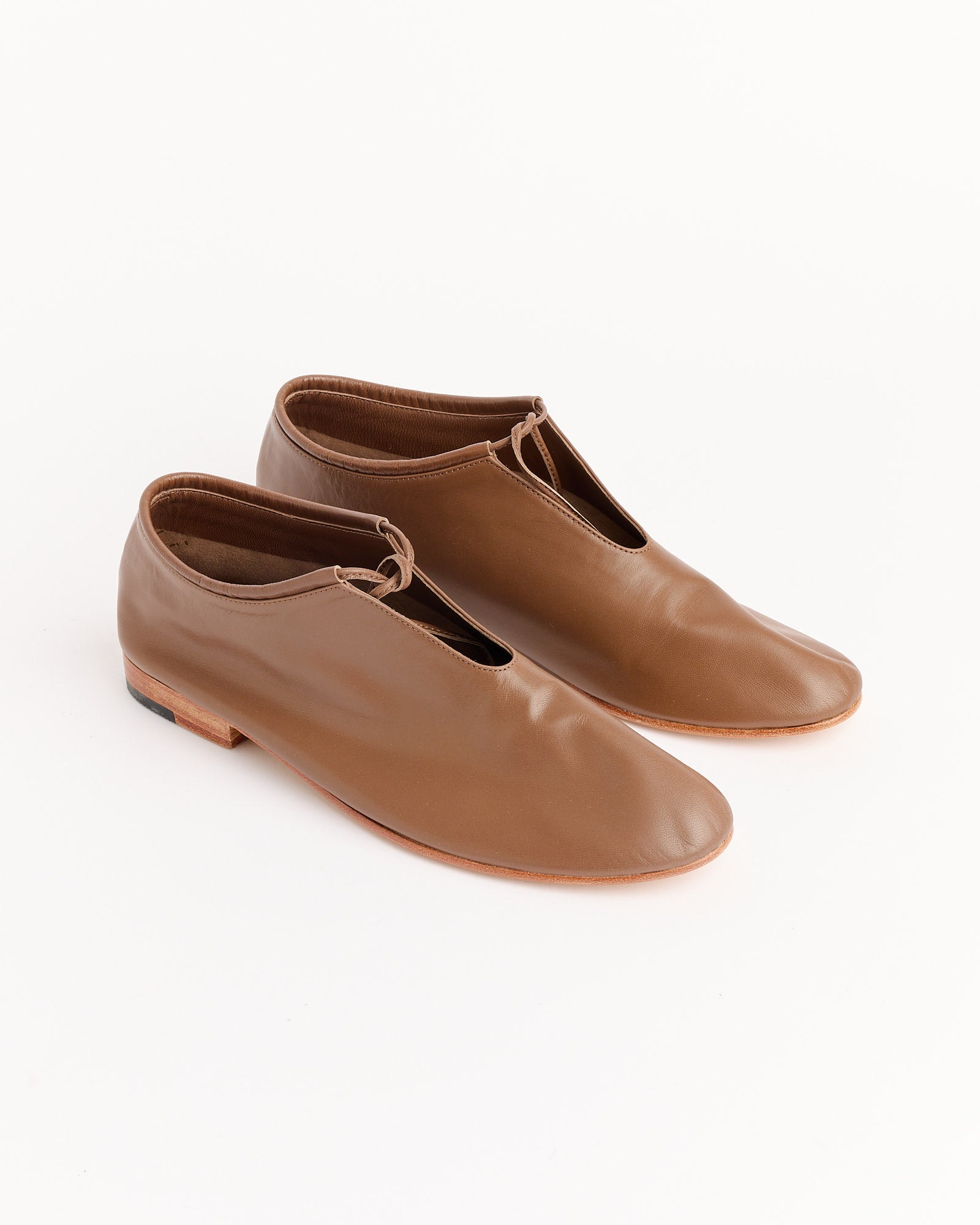 The Martiniano Bootie in Vision features brown handcrafted kid leather slip-on shoes with a small knot detail, flat wooden soles, and a minimalist design, shown on a white background.