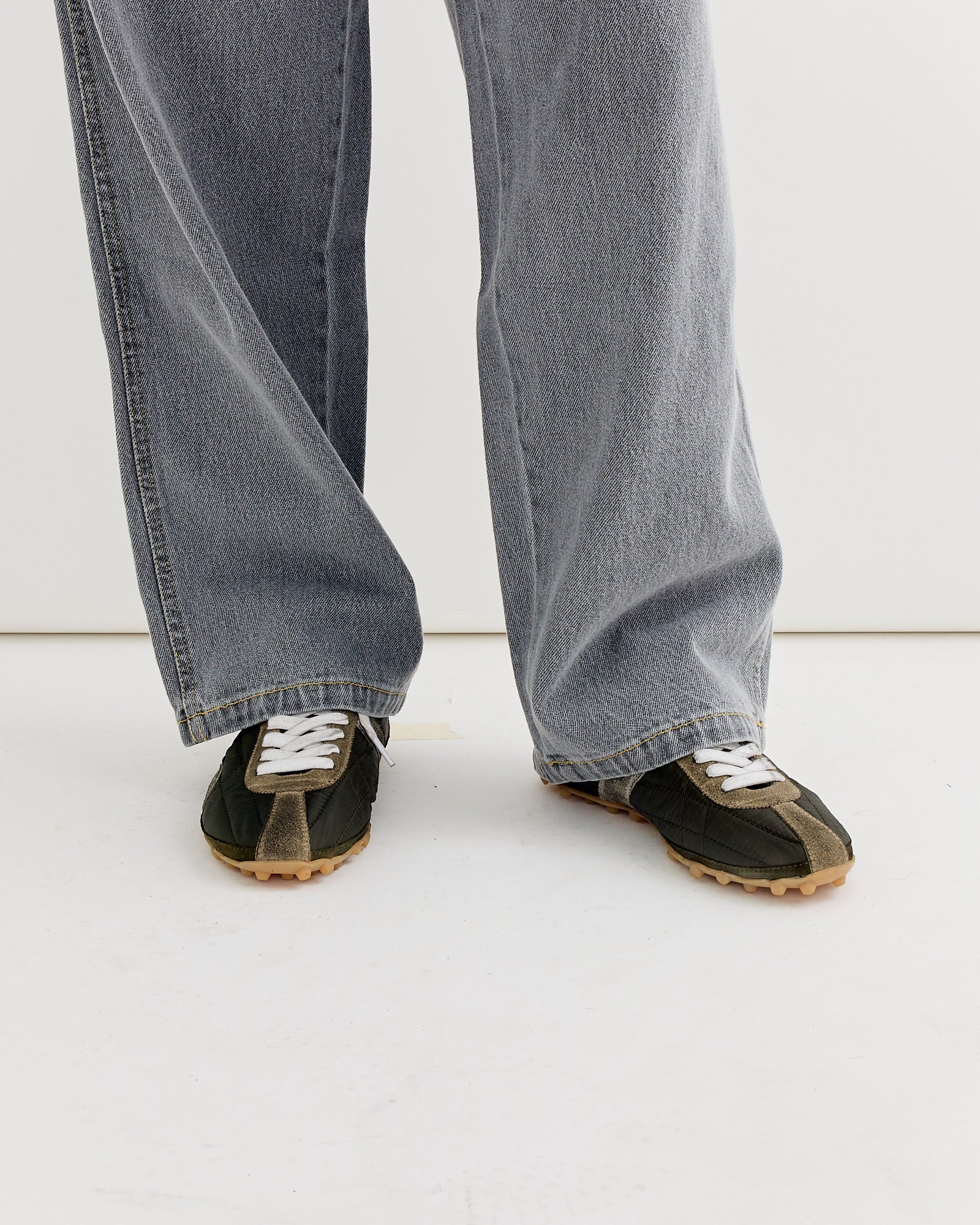 A person wears wide-leg, faded gray jeans with a relaxed fit, partially covering Maison Margiela's Sprinters Low Top in Khaki as they stand on a white floor against a white background, emphasizing the sneakers’ unique sizing.
