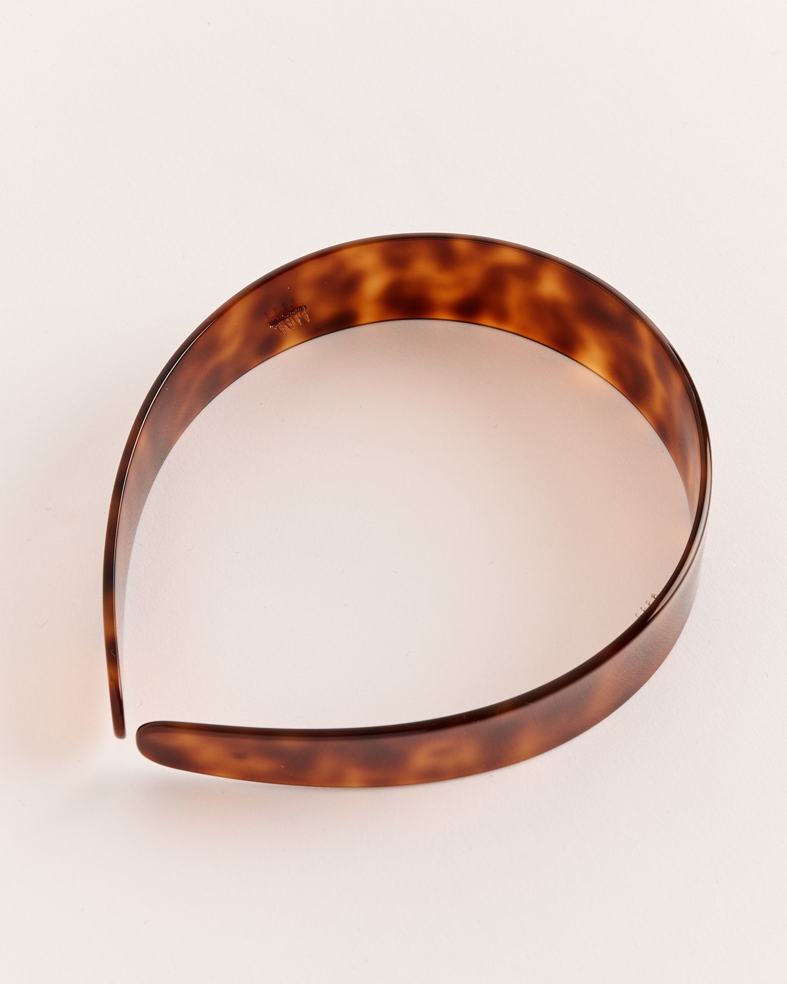 The Thin Bessette Headband in Tortoise by Sophie Buhai, featuring a sleek tortoiseshell pattern and glossy finish, rests on a light, neutral surface.