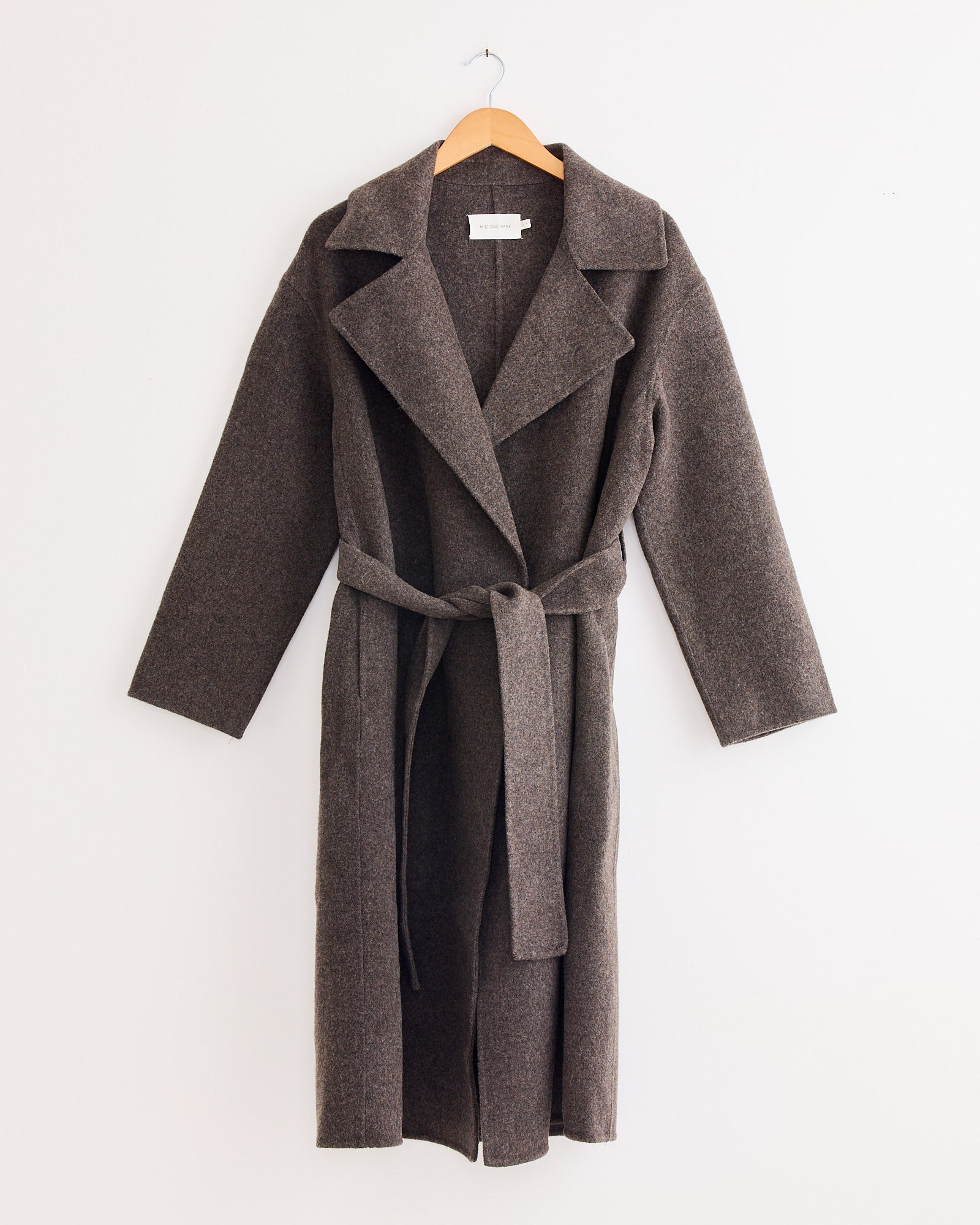 The Handmade Belted Wool Coat in Brown by Mijeong Park hangs on a wooden hanger against a plain white wall, featuring a wide collar and classic belt detail.
