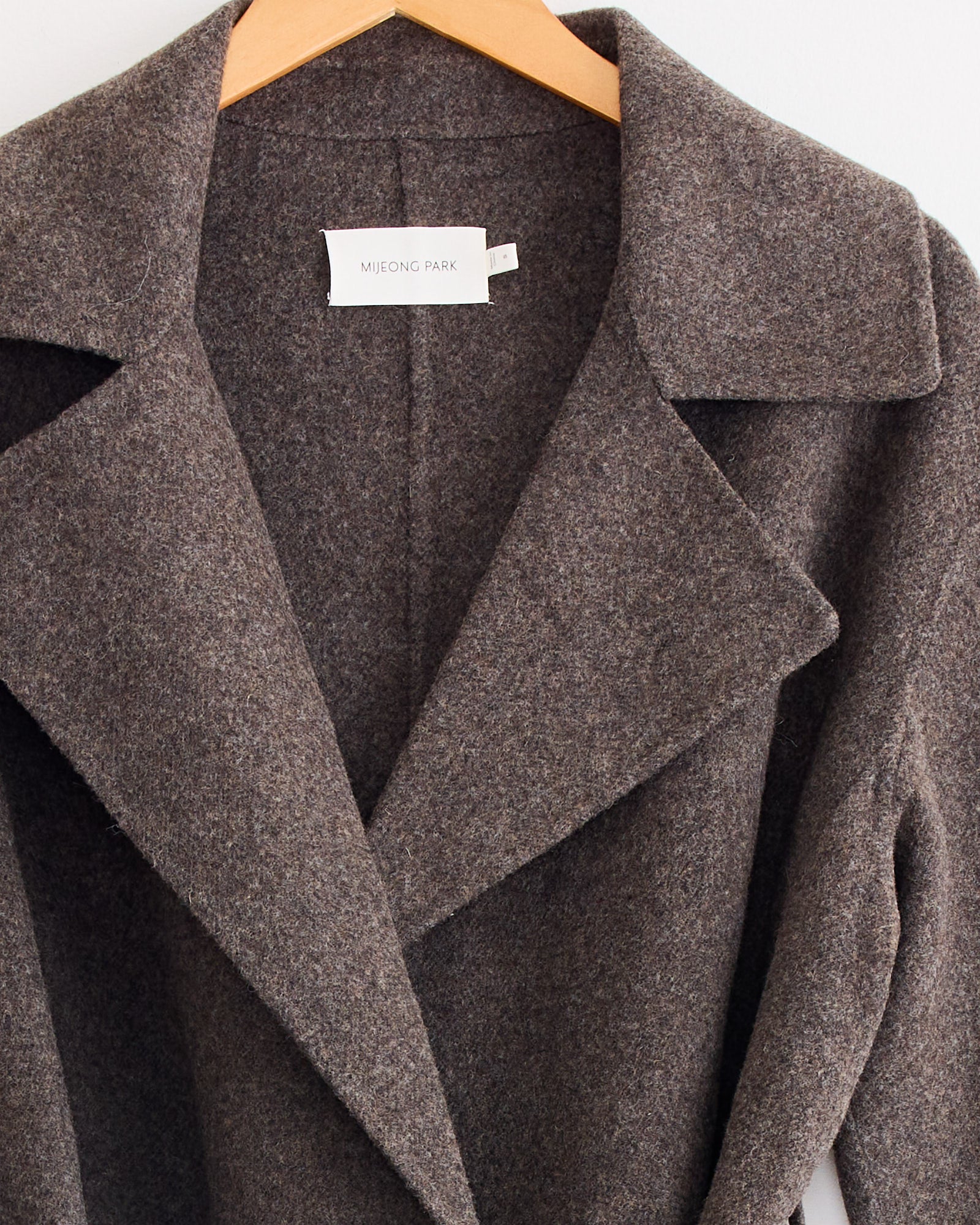 A close-up of the Handmade Belted Wool Coat in Brown by Mijeong Park, featuring wide lapels and hanging on a wooden hanger, with a white Mijeong Park label inside the collar.