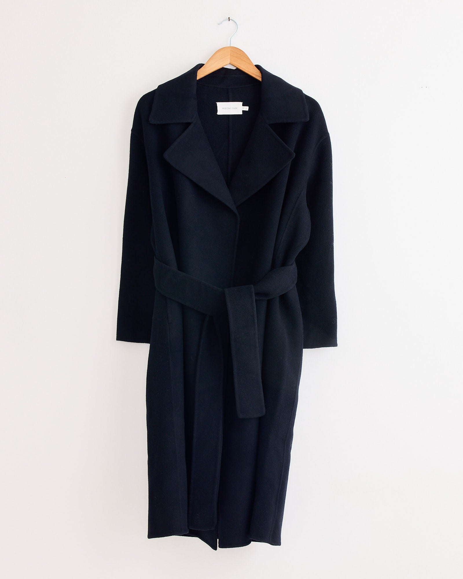 The Mijeong Park Handmade Belted Wool Coat in Navy features a wide collar and matching fabric belt, hanging elegantly on a wooden hanger against a plain white wall.