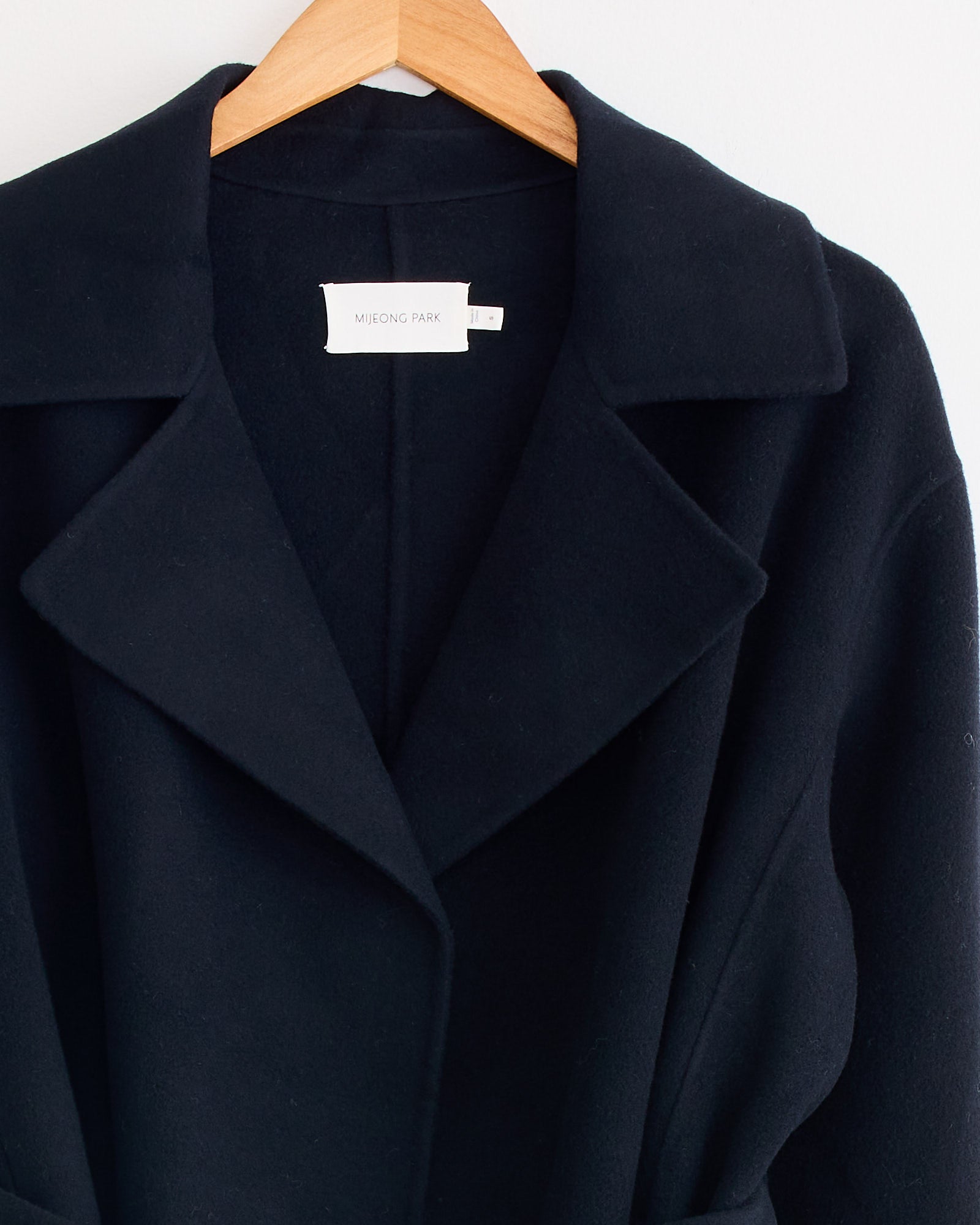 A Handmade Belted Wool Coat in Navy by Mijeong Park hangs on a wooden hanger, featuring wide lapels and a white brand label inside the collar, set against a plain white wall.