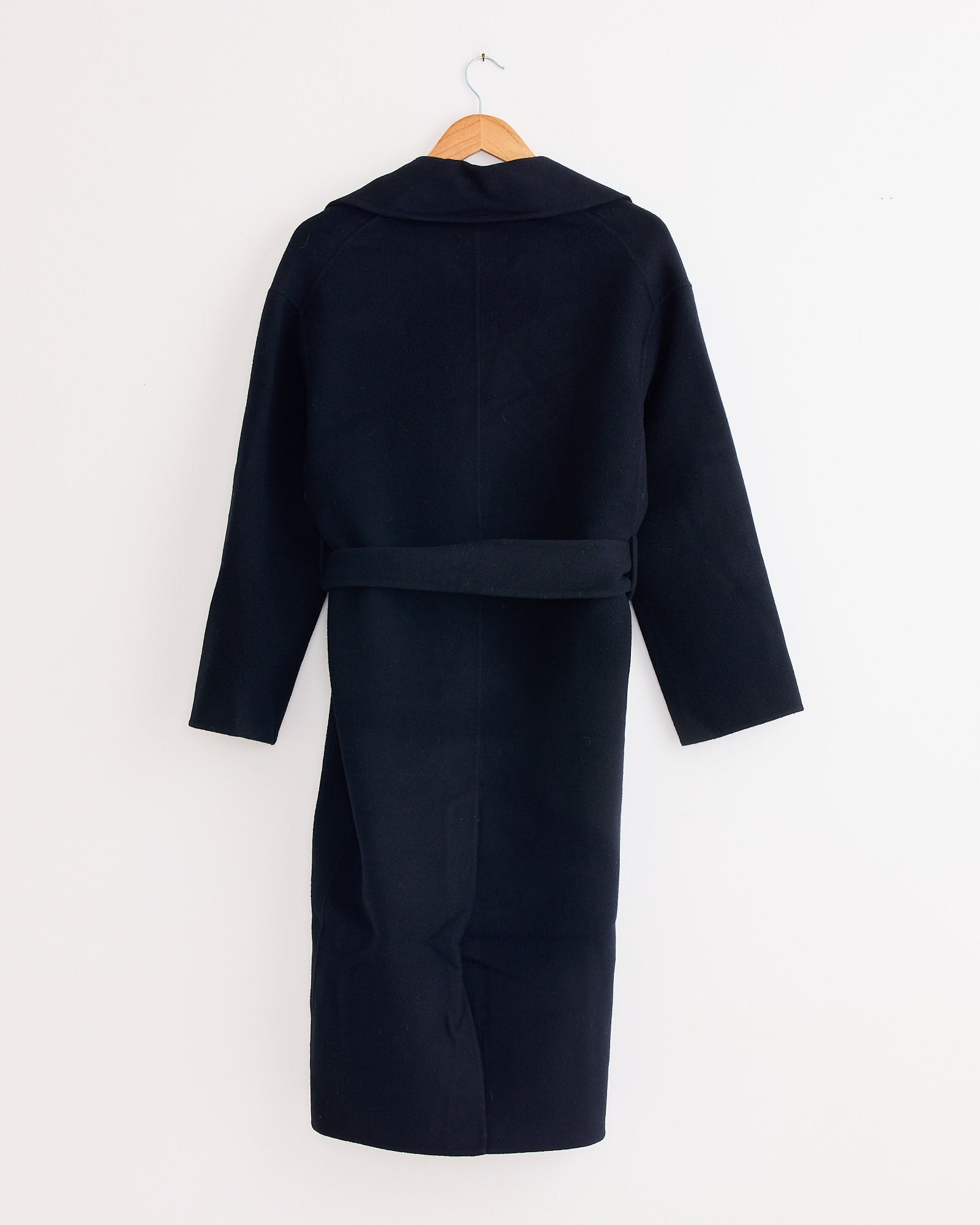 The Handmade Belted Wool Coat in Navy by Mijeong Park hangs on a wooden hanger against a plain white wall, displayed from the back for a timeless, understated look.