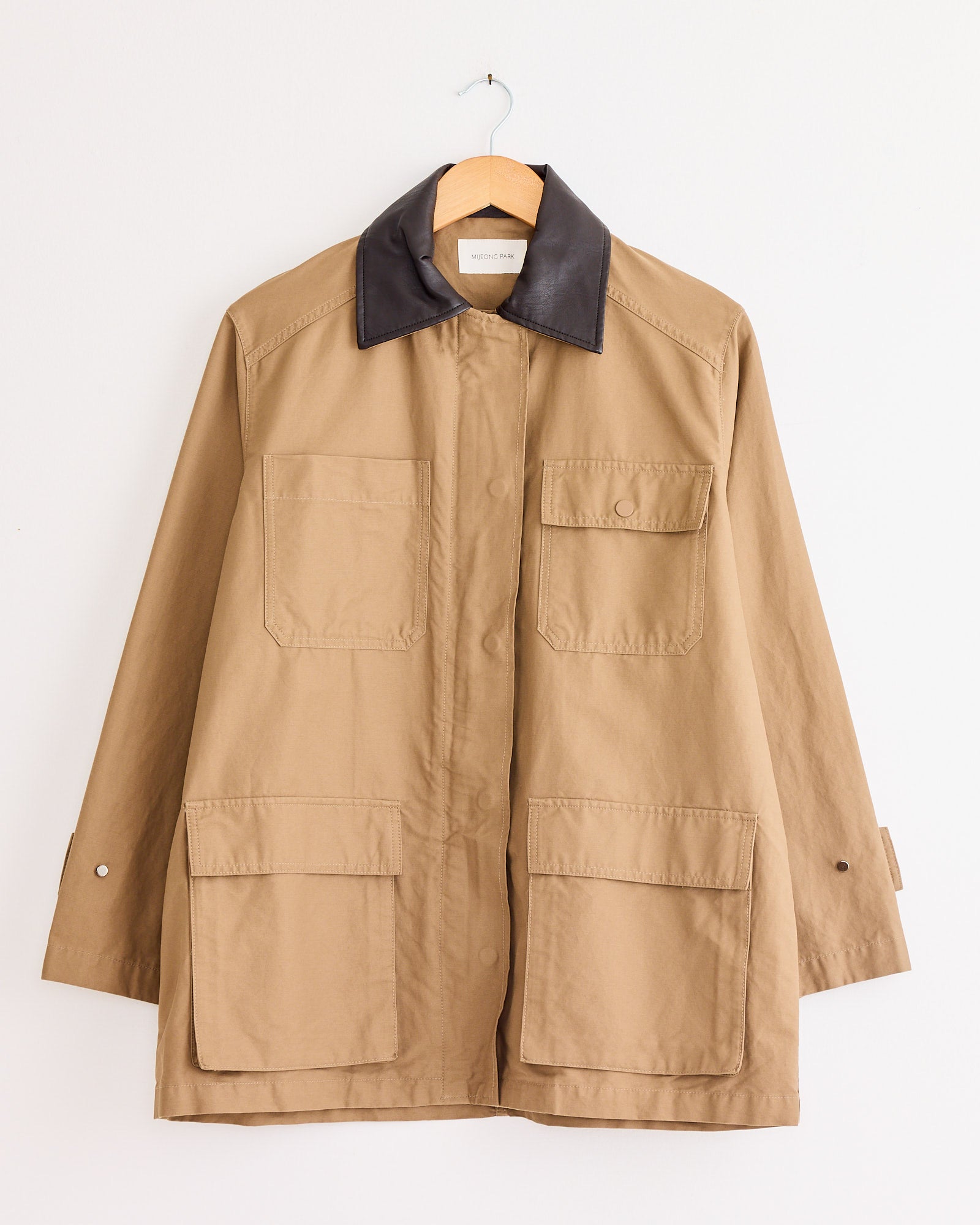 The Mijeong Park Contrast Collar Work Jacket in Dark Beige features a dark brown collar, four large front pockets, and buttoned cuffs. It hangs on a wooden hanger against a plain white wall.