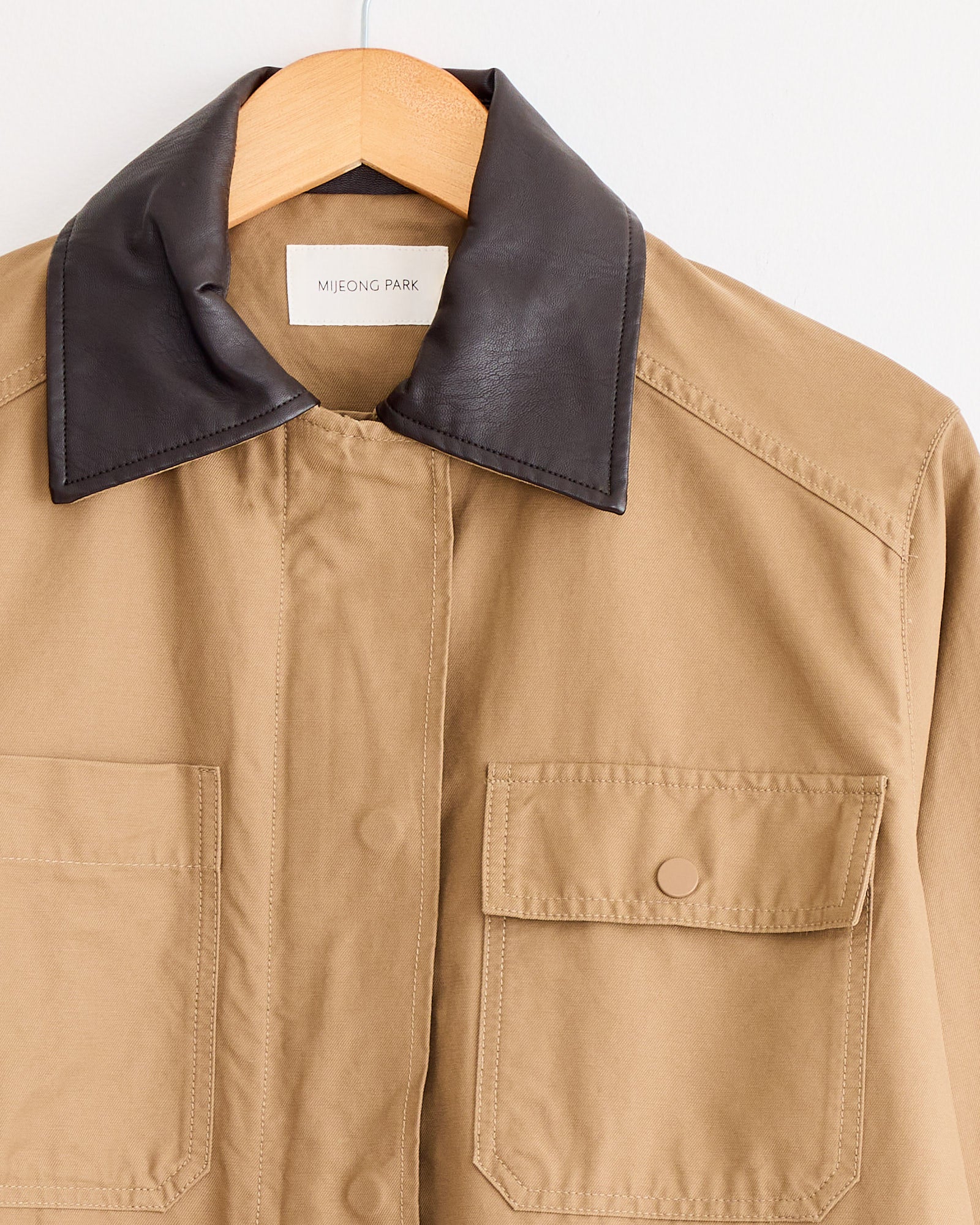 The Mijeong Park Contrast Collar Work Jacket in Dark Beige, featuring a dark brown leather collar and front flap pockets, hangs neatly on a wooden hanger.