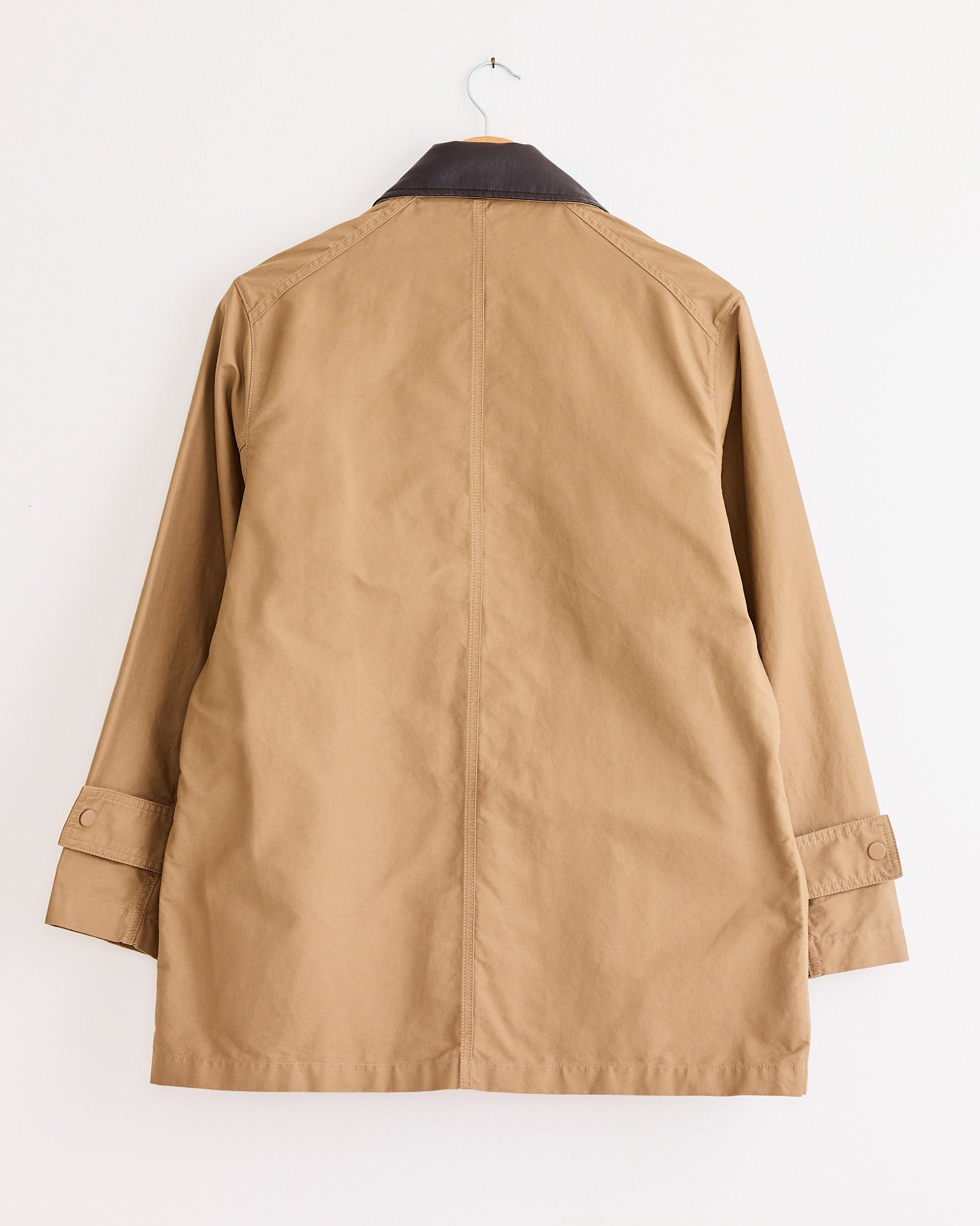 The Mijeong Park Contrast Collar Work Jacket in Dark Beige features a dark brown trim, long button-cuffed sleeves, and a center seam for structure. This oversized cotton jacket is shown hanging from the back on a wall-facing hook.