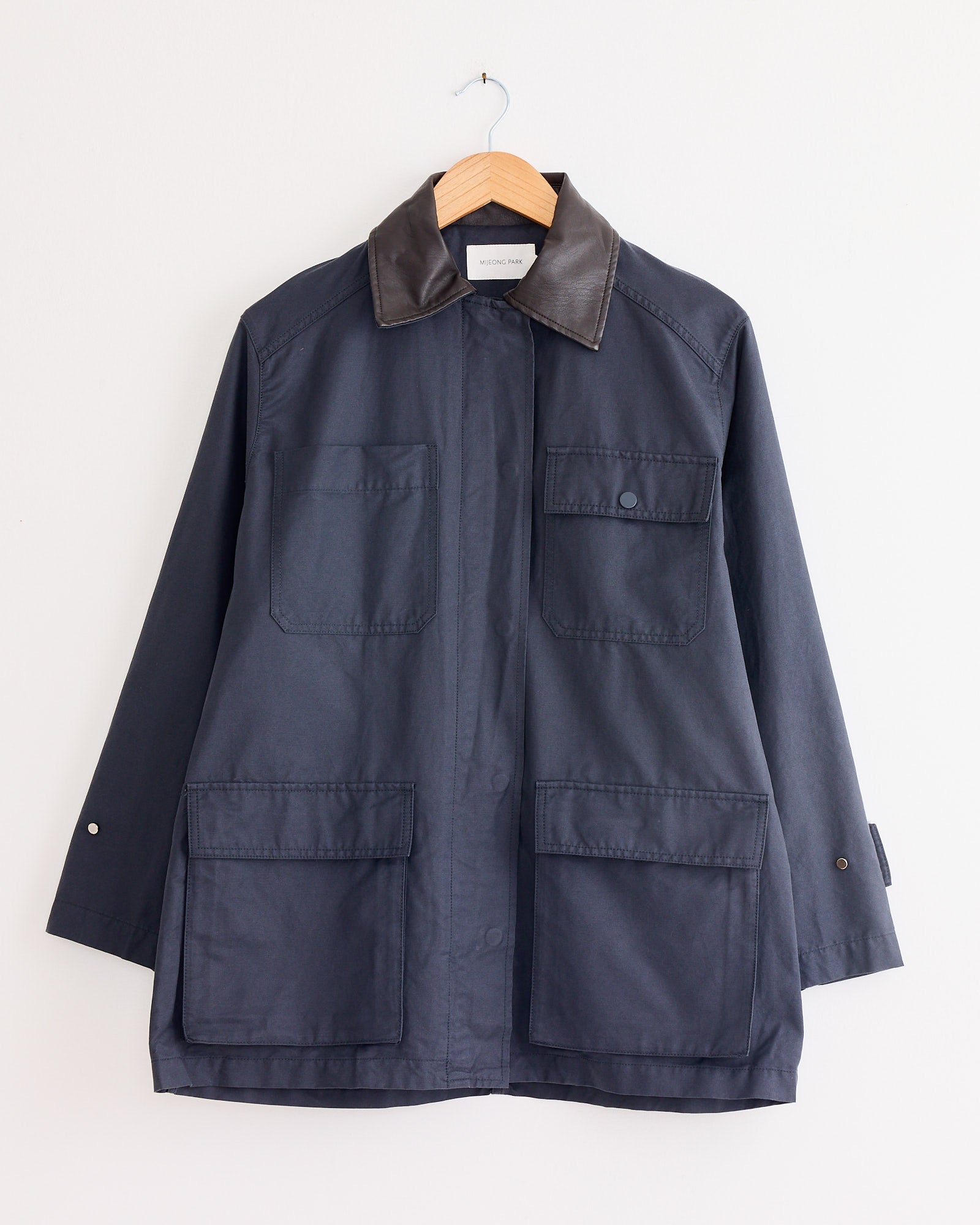 The Contrast Collar Work Jacket in Navy by Mijeong Park, with a brown collar, buttoned front, and four large pockets, is displayed on a wooden hanger against a white wall.
