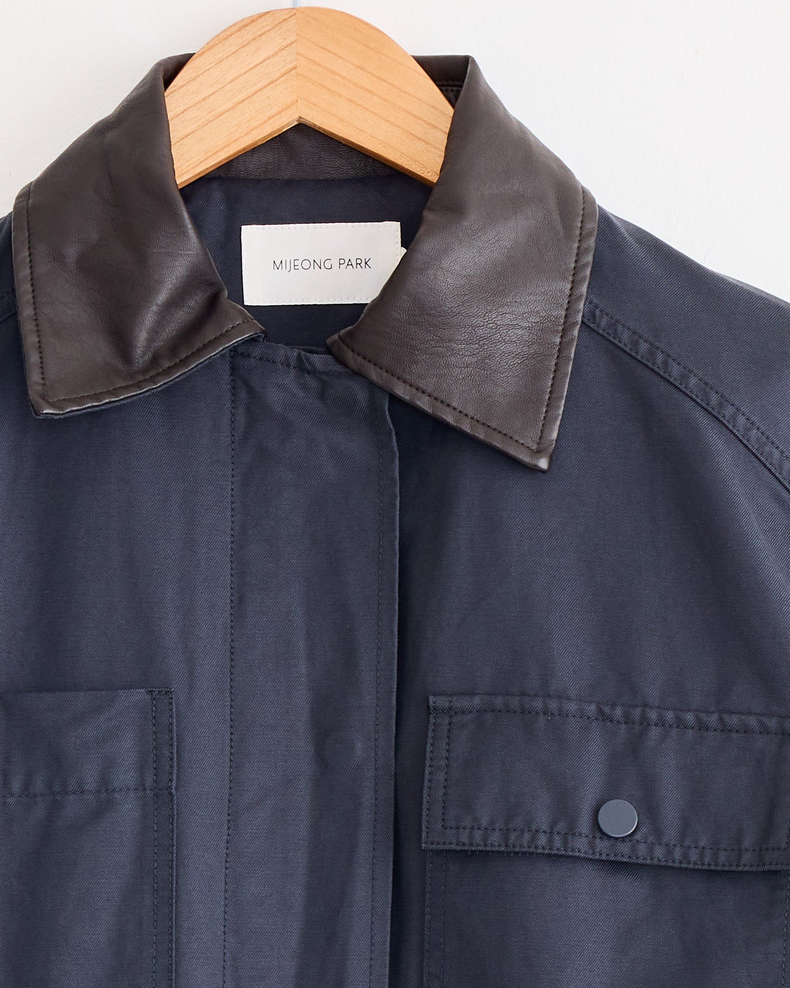 The Mijeong Park Contrast Collar Work Jacket in Navy hangs on a wooden hanger, featuring a black leather collar, oversized cotton fit, front flap pockets with snap buttons, and a signature label inside.