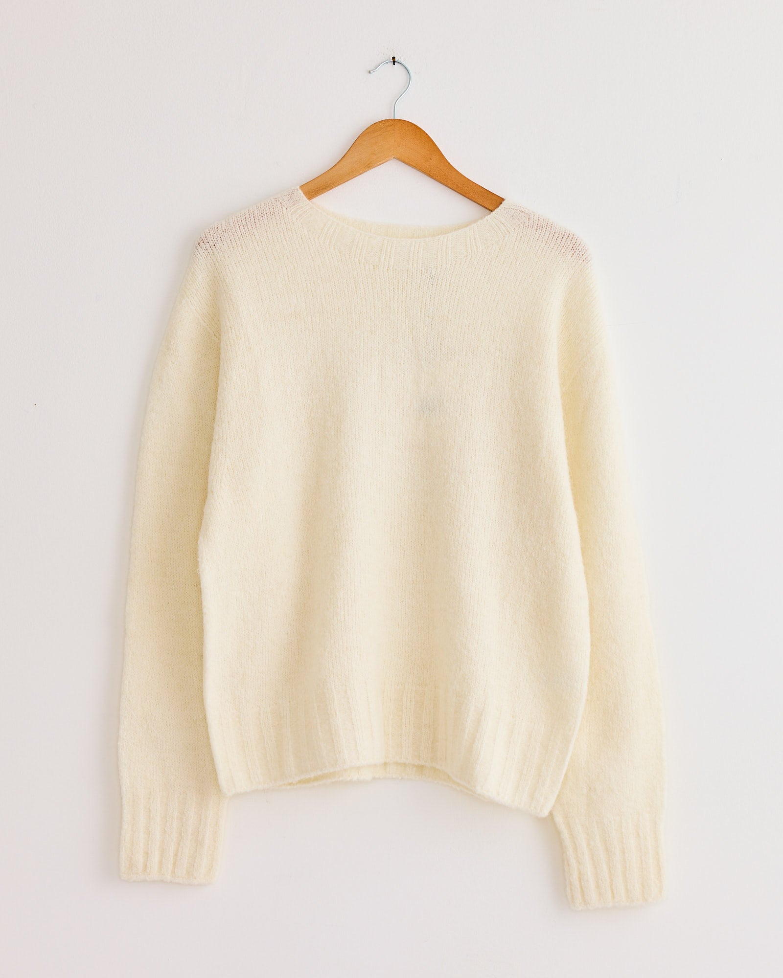 The Mijeong Park Alpaca Blend Wholegarment Knit in Ivory, an oversized cream-colored sweater, hangs on a wooden hanger against a plain white wall.