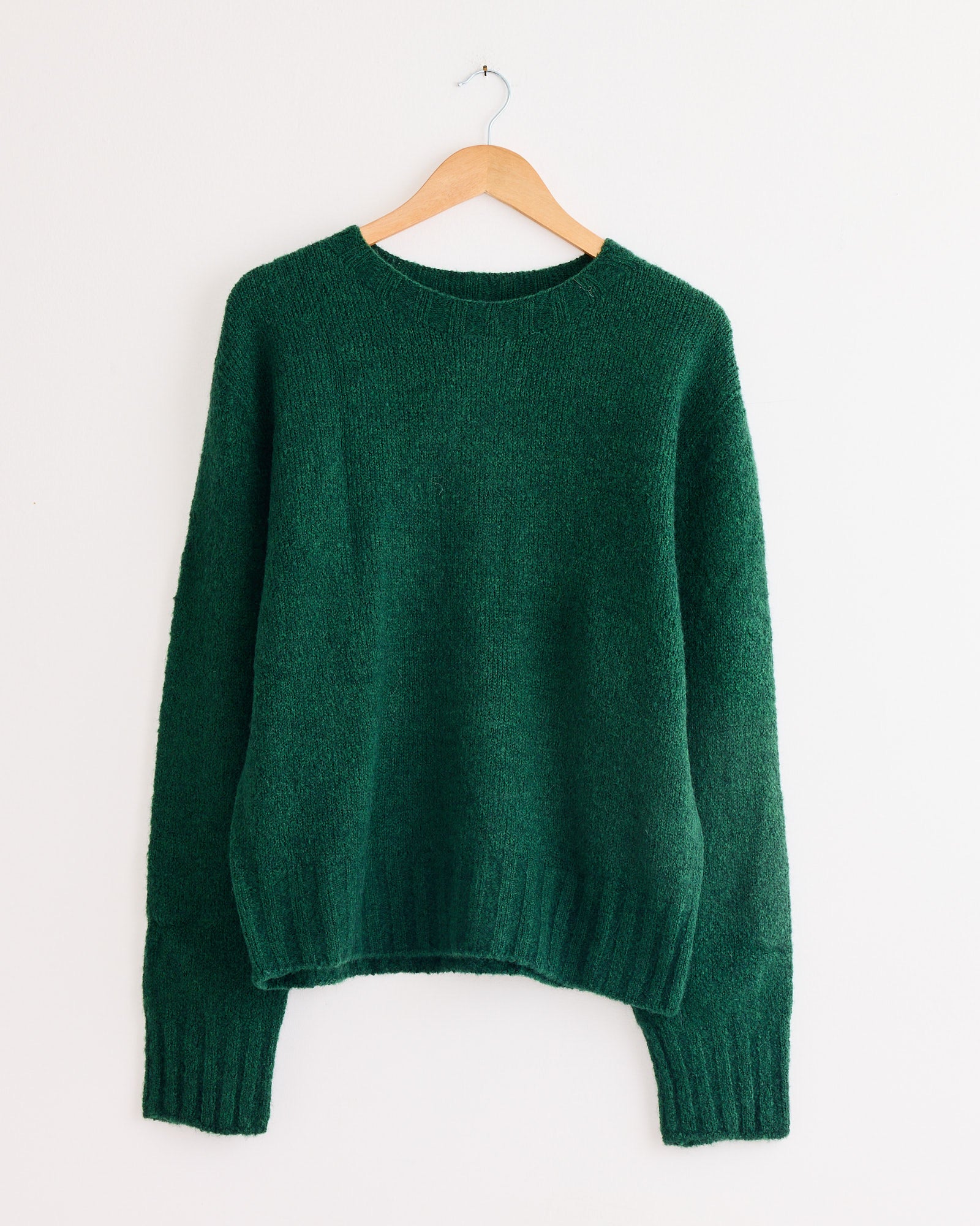 The Mijeong Park Alpaca Blend Wholegarment Knit in green, featuring an oversized fit and long sleeves, hangs on a wooden hanger against a plain white wall.