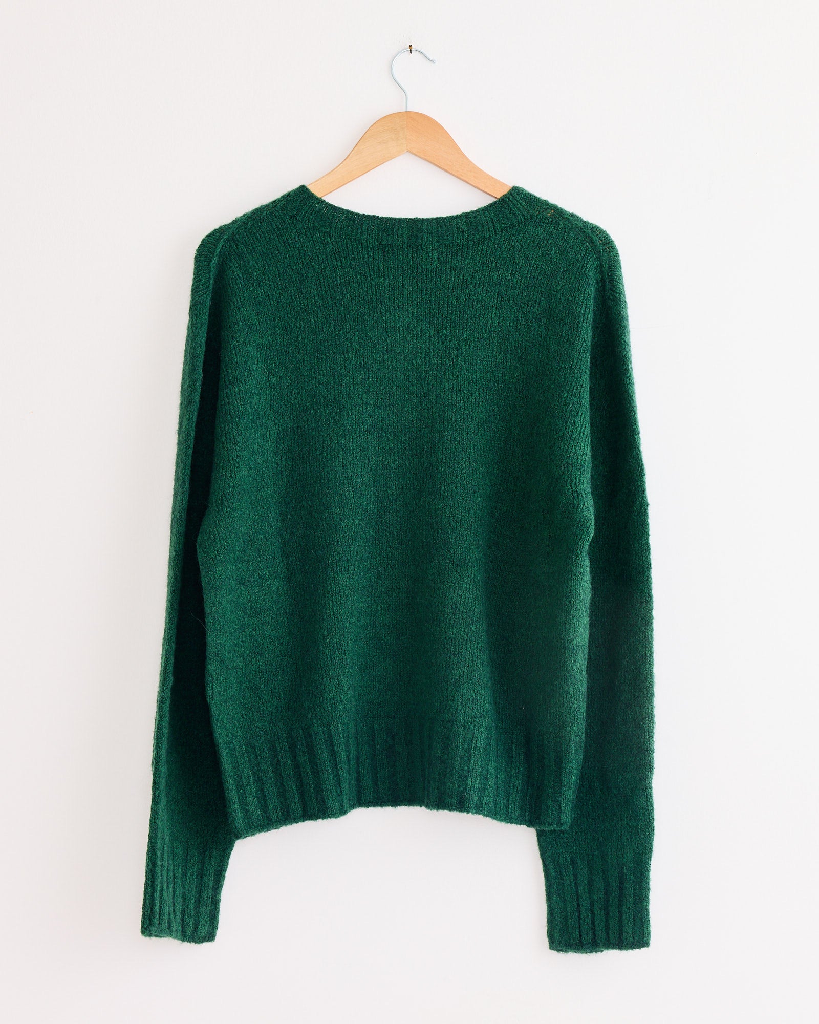 The Mijeong Park Alpaca Blend Wholegarment Knit in Green, an oversized sweater with long sleeves, hangs on a wooden hanger against a plain white wall.