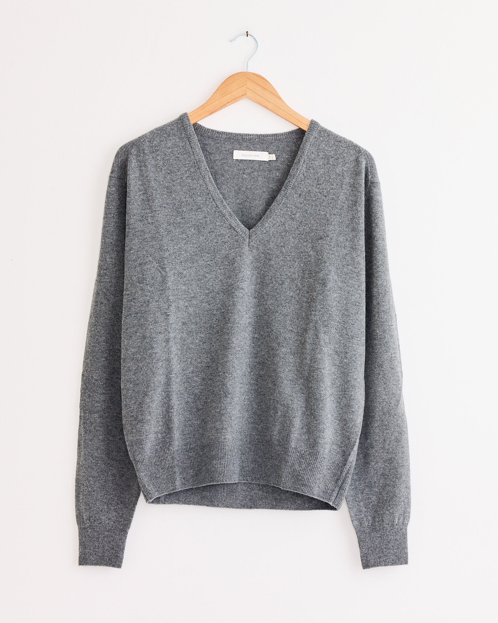 The Mijeong Park Cashmere Blend V-Neck Knit in Grey, with long sleeves, hangs on a wooden hanger against a white wall, highlighting its soft texture.