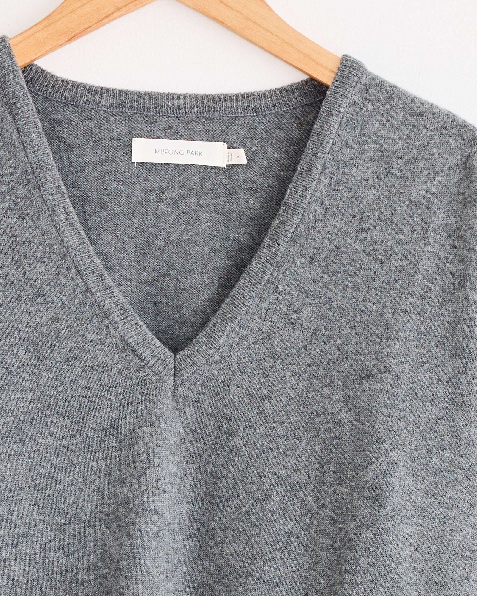 A close-up of the Mijeong Park Cashmere Blend V-Neck Knit in Grey on a wooden hanger, featuring a white label with the brand name inside the collar.
