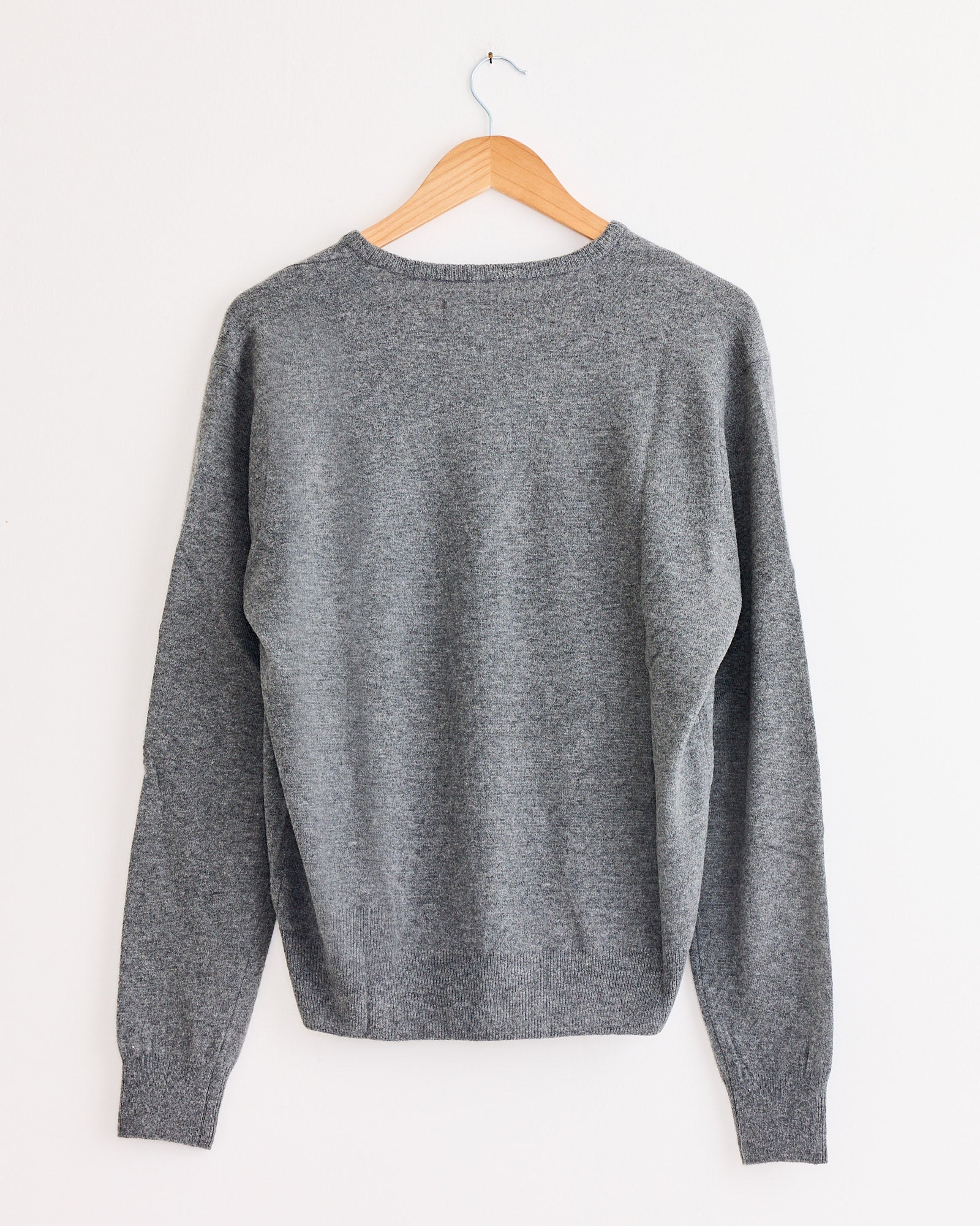 The Mijeong Park Cashmere Blend V-Neck Knit in Grey hangs on a wooden hanger against a white wall, displayed from the back with simple ribbed cuffs and hem for a timeless look.