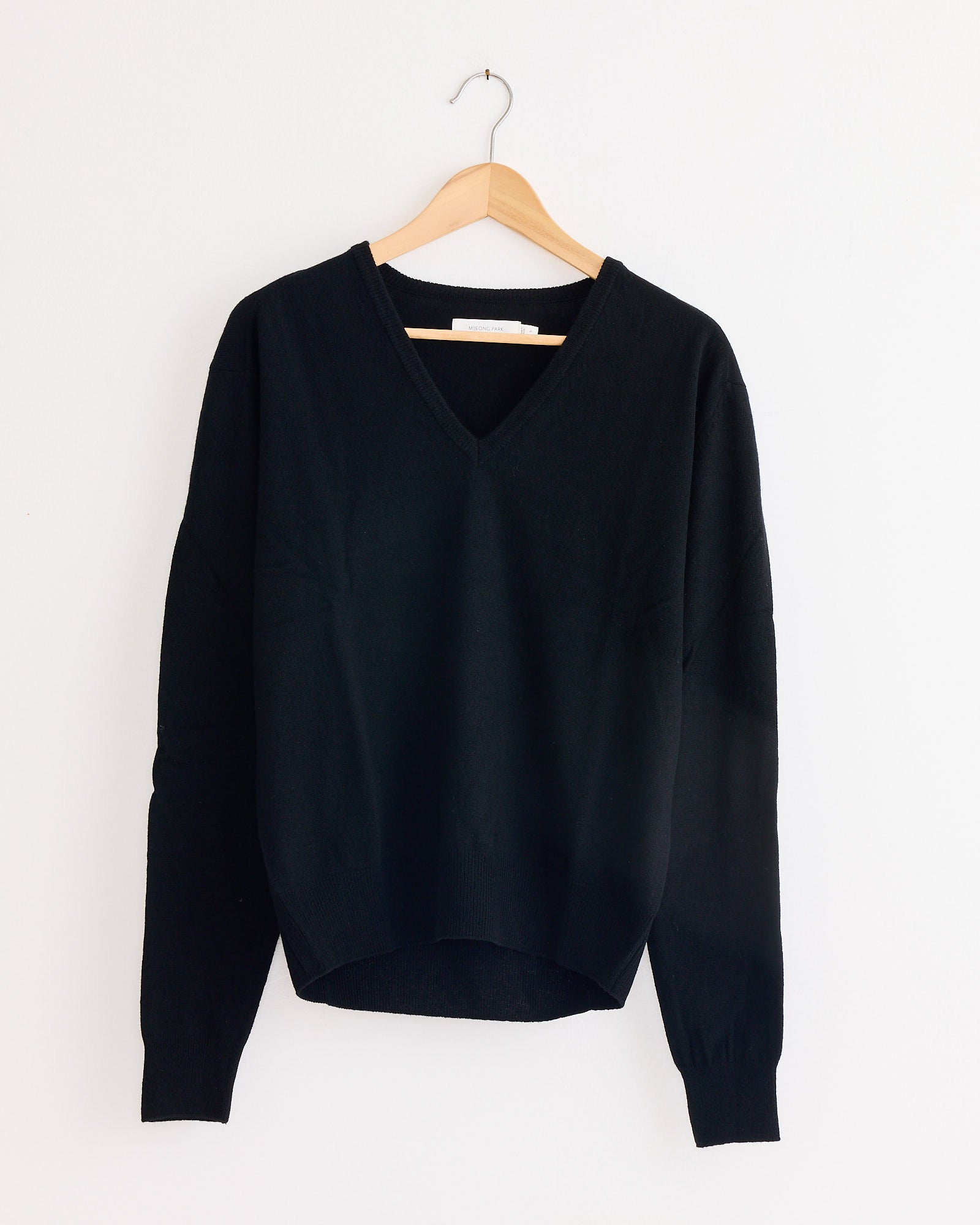 The Mijeong Park Cashmere Blend V-Neck Knit in Black hangs on a wooden hanger against a plain white wall, highlighting its classic long-sleeve design.