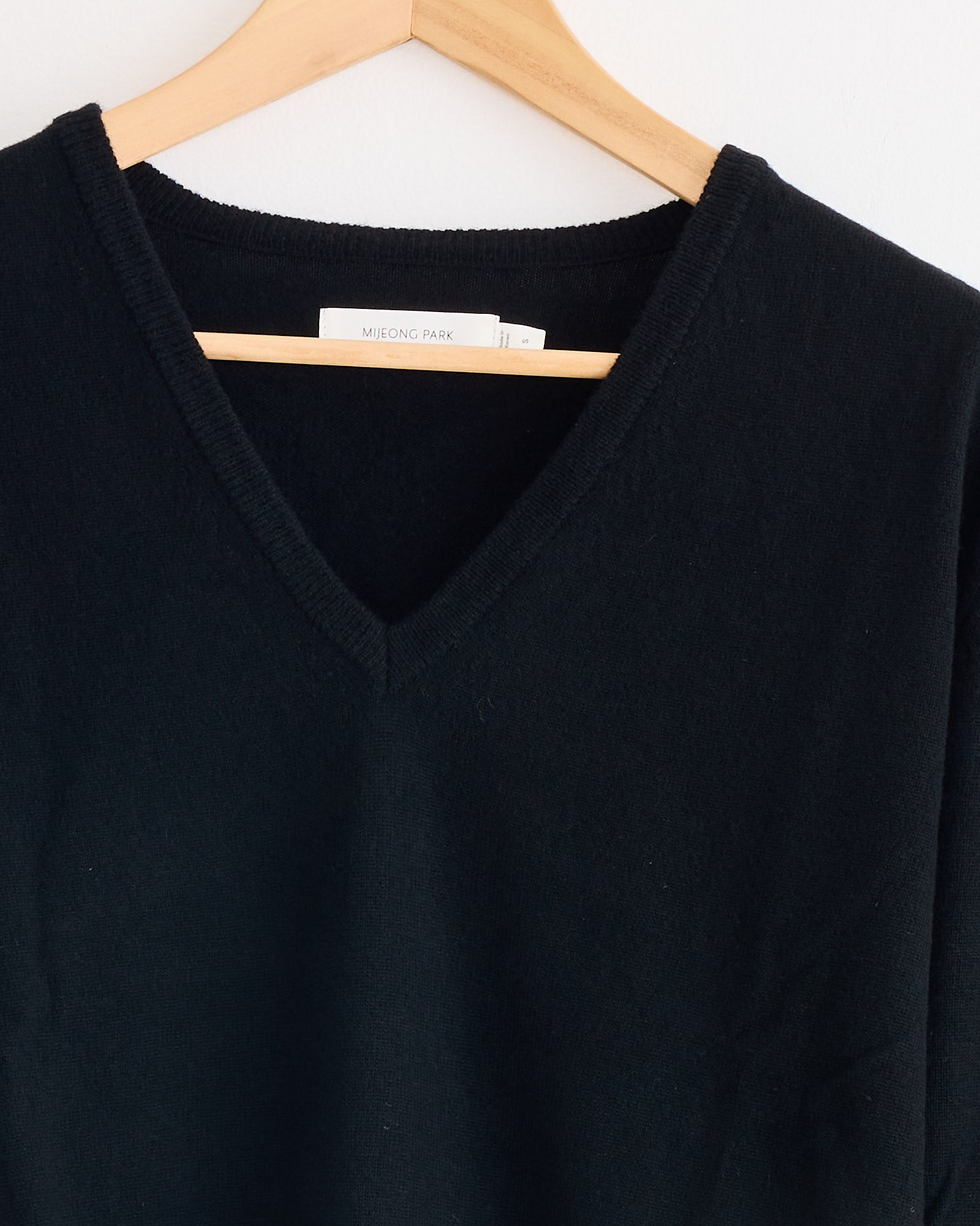 A black Mijeong Park Cashmere Blend V-Neck Knit hangs on a wooden hanger against a white background, featuring a label inside the collar.