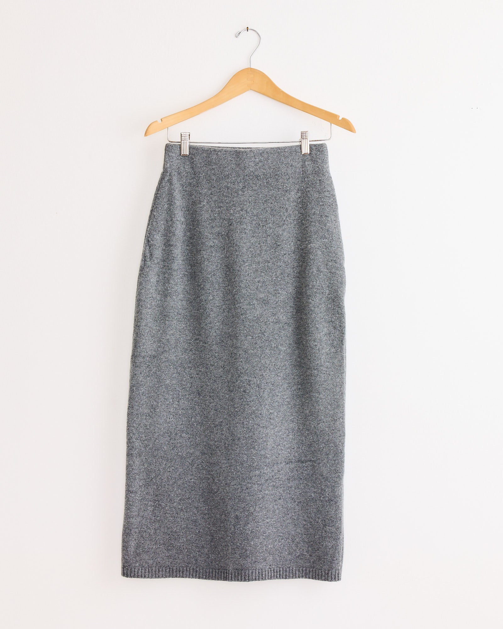 The Mijeong Park Cashmere Blend Knit Midi Skirt in Grey, with a simple straight design, hangs on a wooden hanger against a plain white wall.