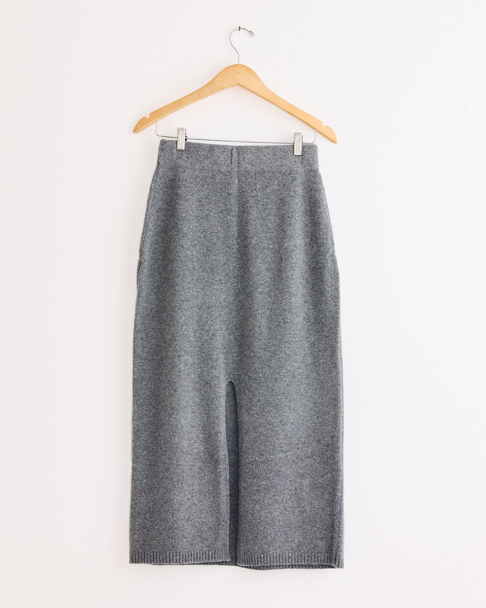 The Mijeong Park Cashmere Blend Knit Midi Skirt in Grey hangs on a wooden hanger against a white wall, featuring a straight cut and ribbed hem with no visible patterns or embellishments.