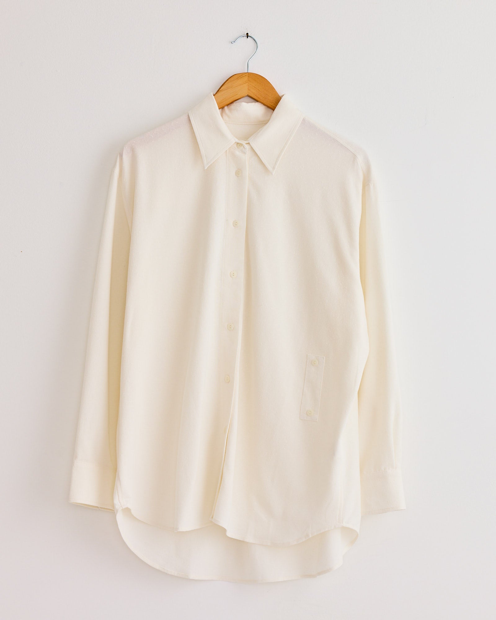 The Mijeong Park Textured Oversized Shirt in Ivory features a classic collar, long sleeves, button-up front, and relaxed fit, crafted from a textured cotton blend. It hangs on a wooden hanger against a plain white wall.