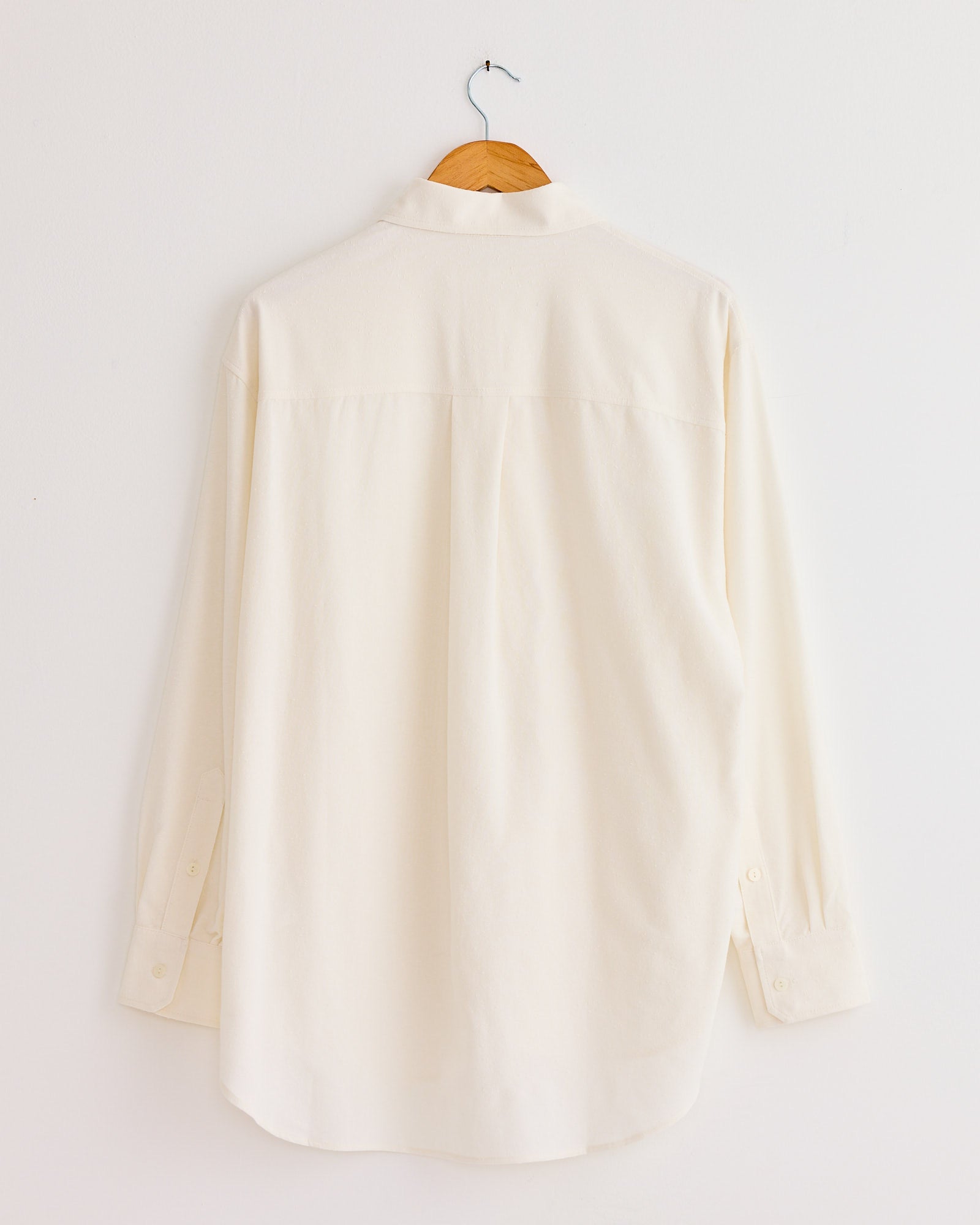 The Mijeong Park Textured Oversized Shirt in Ivory, featuring long sleeves and a cotton blend, hangs on a wooden hanger against a plain white wall, shown from the back.