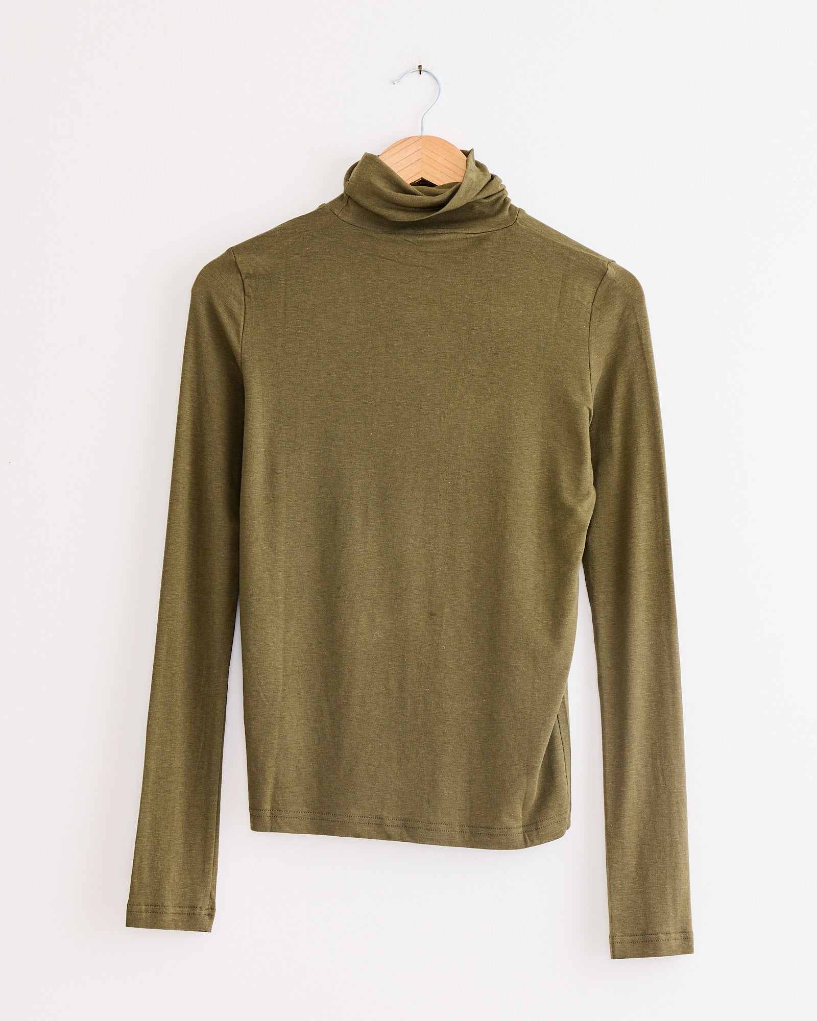 The Mijeong Park Roll Neck Jersey Top Light in Olive, featuring long sleeves and crafted from a lightweight jersey blend, is displayed on a wooden hanger against a plain white wall.