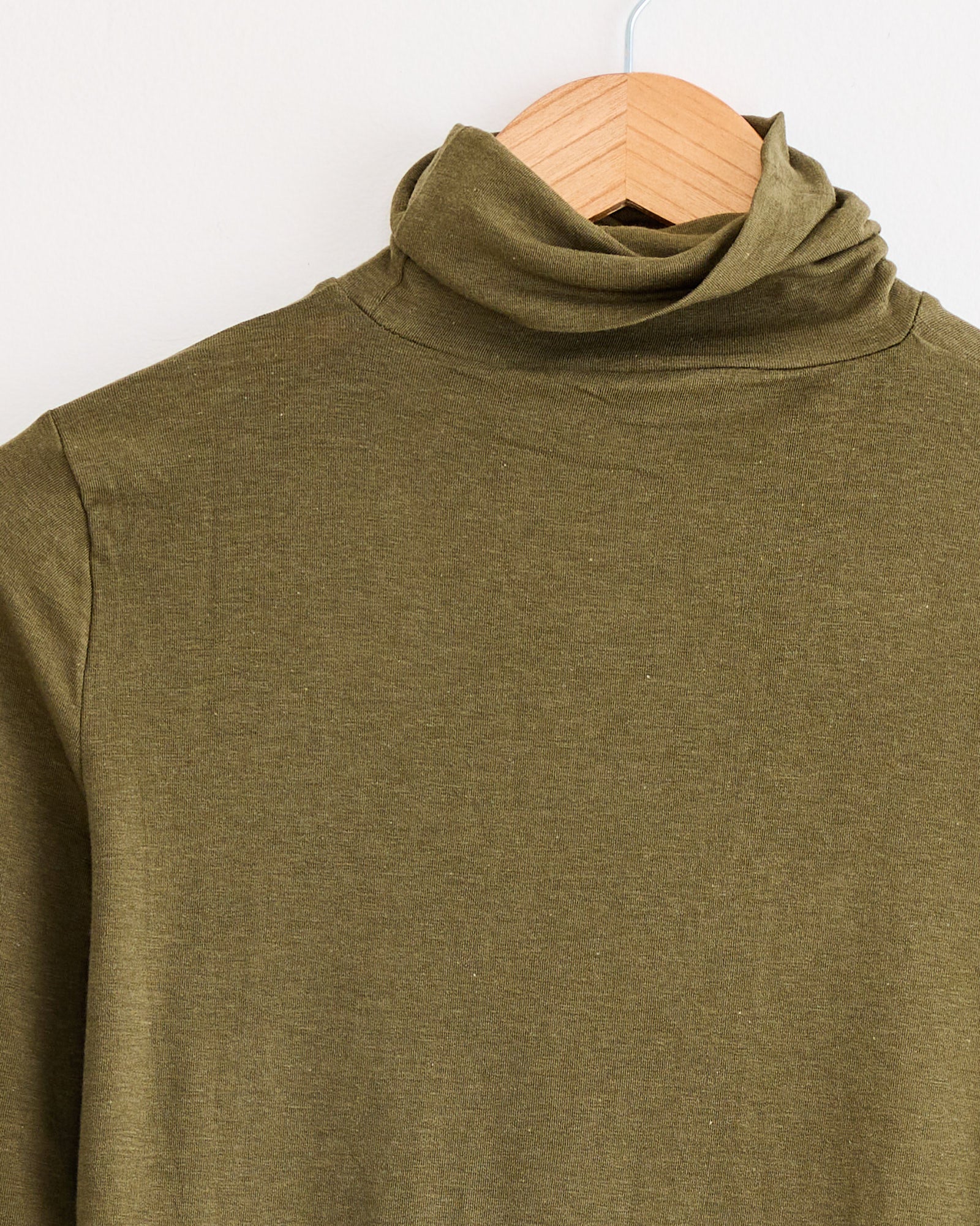The Mijeong Park Roll Neck Jersey Top Light in Olive features a slim fit and folded collar, crafted from a lightweight jersey blend and displayed on a wooden hanger against a white wall.
