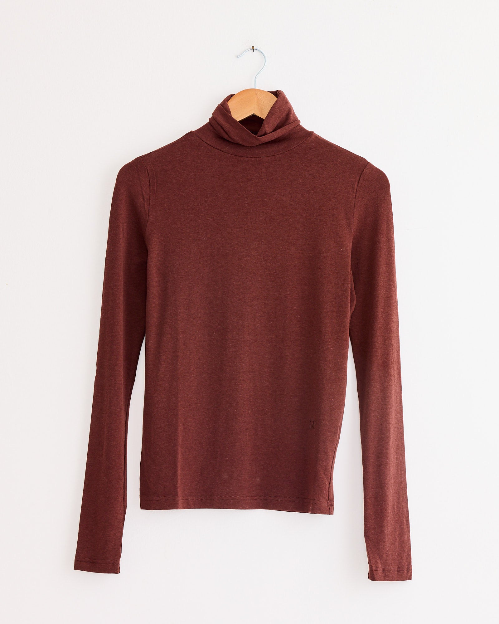 The Mijeong Park Roll Neck Jersey Top in Burgundy, crafted from a lightweight jersey blend, features long sleeves and hangs on a wooden hanger against a plain white wall.