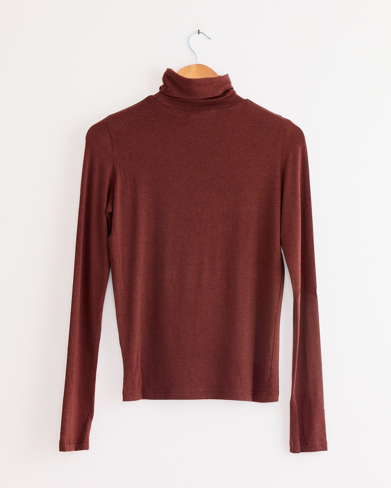 The Mijeong Park Roll Neck Jersey Top in Burgundy, featuring long sleeves and a lightweight jersey blend, hangs on a wooden hanger against a plain white wall.
