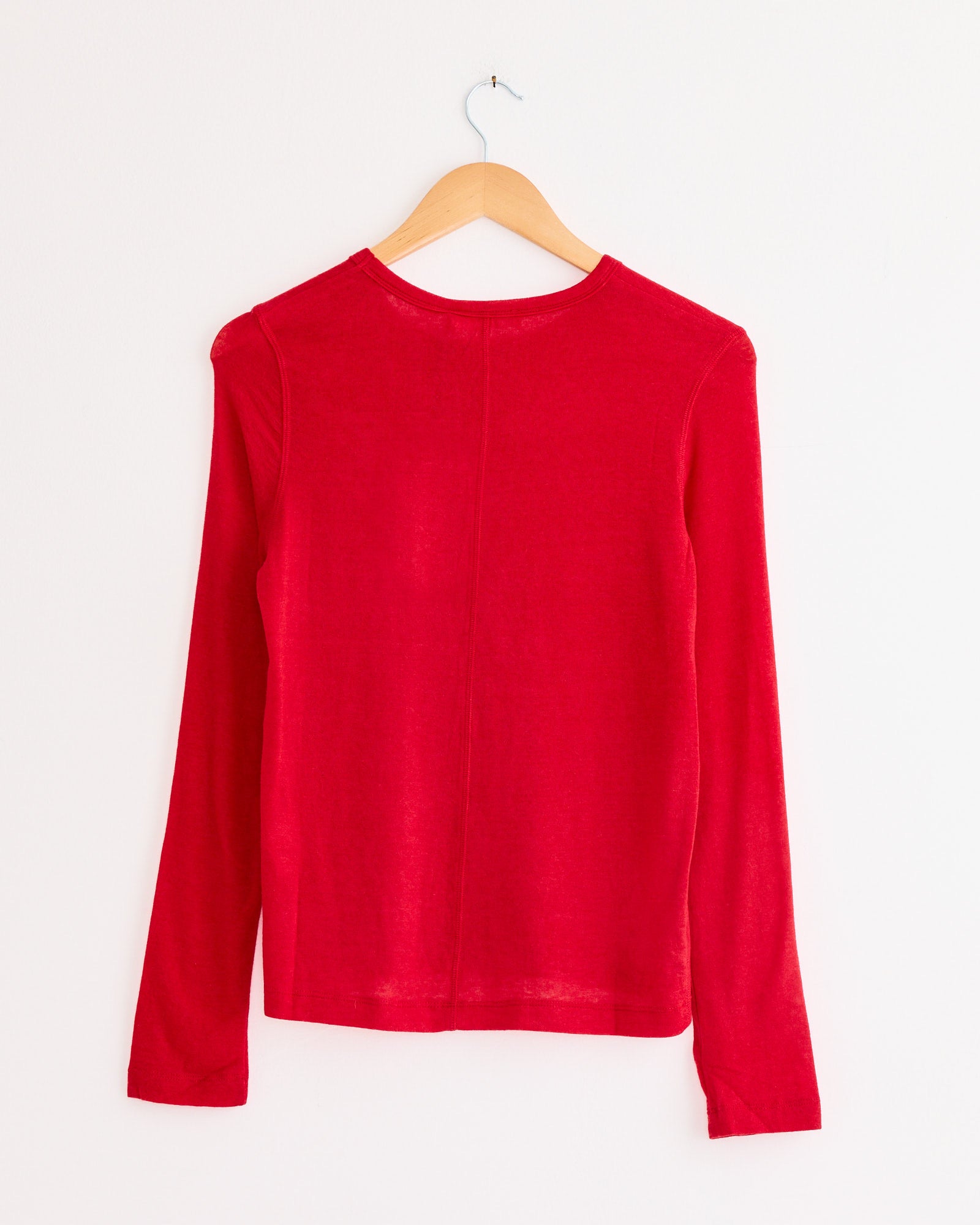 The Mijeong Park Wool Tencel Crew Neck Top in Red, featuring long sleeves and a plain design, hangs on a wooden hanger with the back facing forward against a white wall.