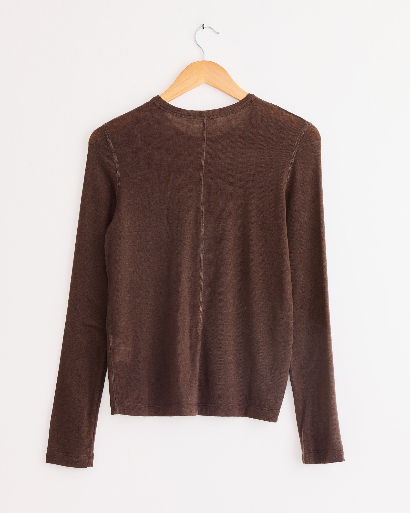 The Mijeong Park Wool Tencel Crew Neck Top in Brown, featuring a slim fit and long sleeves, is displayed on a wooden hanger against a white wall, with the back of the shirt facing forward.