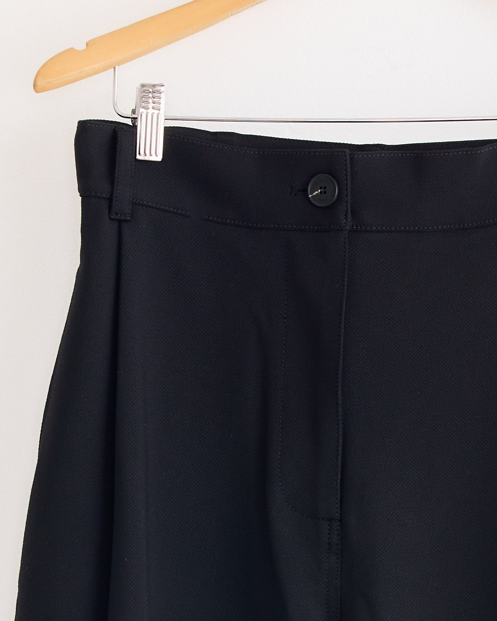 Close-up of the Mijeong Park Pleated Barrel Leg Pant in Black hanging on a wooden hanger, highlighting the adjustable waistband, button, zipper, and a metal clip.