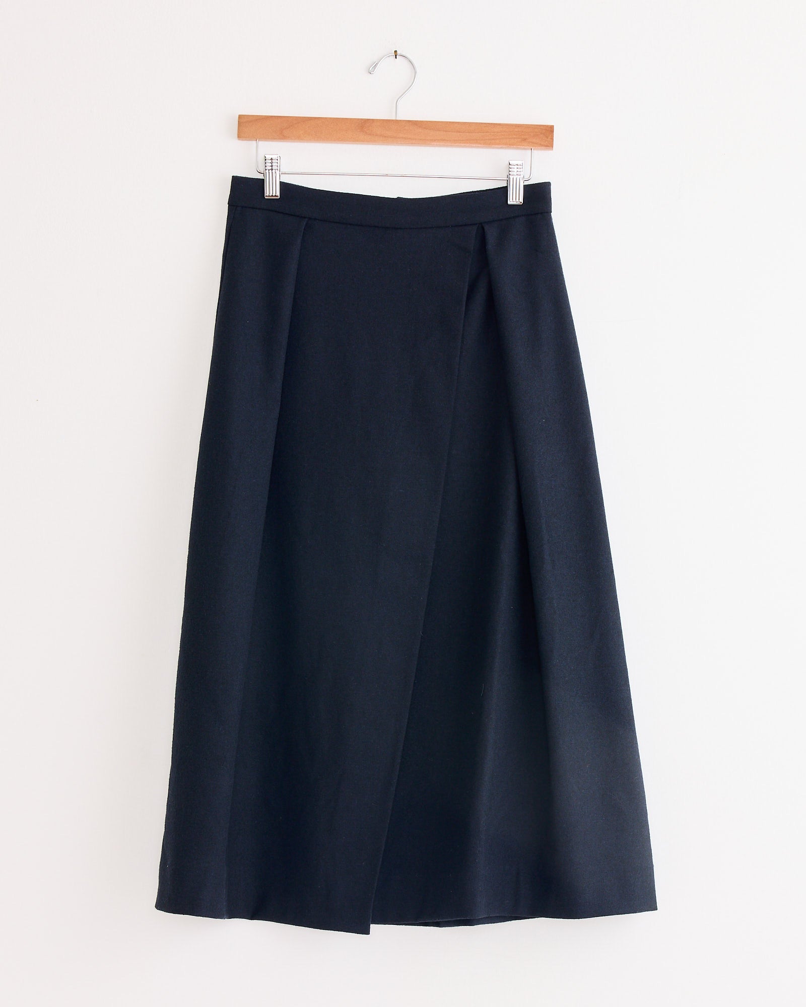 The Mijeong Park Wool Blend Pleated Midi Skirt in Navy, with a subtle front overlap, hangs on a wooden hanger against a plain white background.