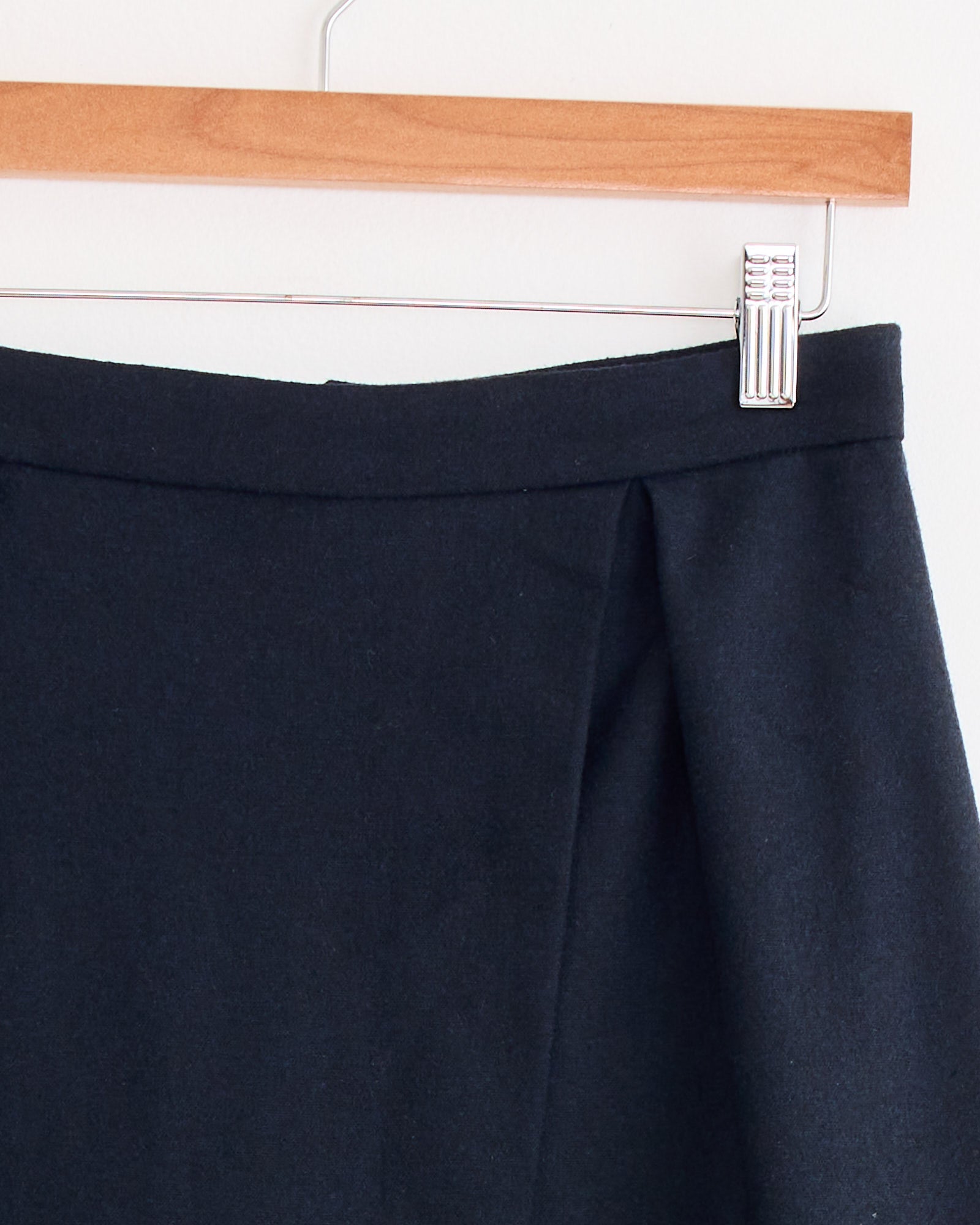 Close-up of the Mijeong Park Wool Blend Pleated Midi Skirt in Navy, showcasing its pleat detail as it hangs on a wooden hanger against a light-colored wall.