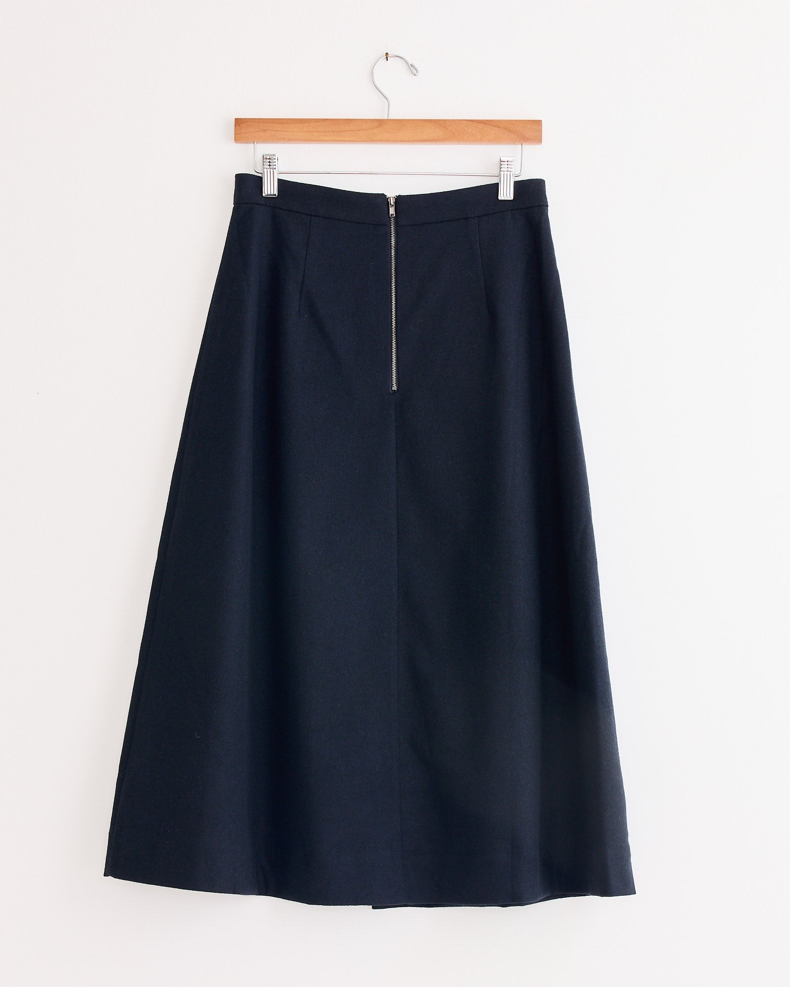 The Mijeong Park Wool Blend Pleated Midi Skirt in Navy hangs on a wooden hanger against a white wall.