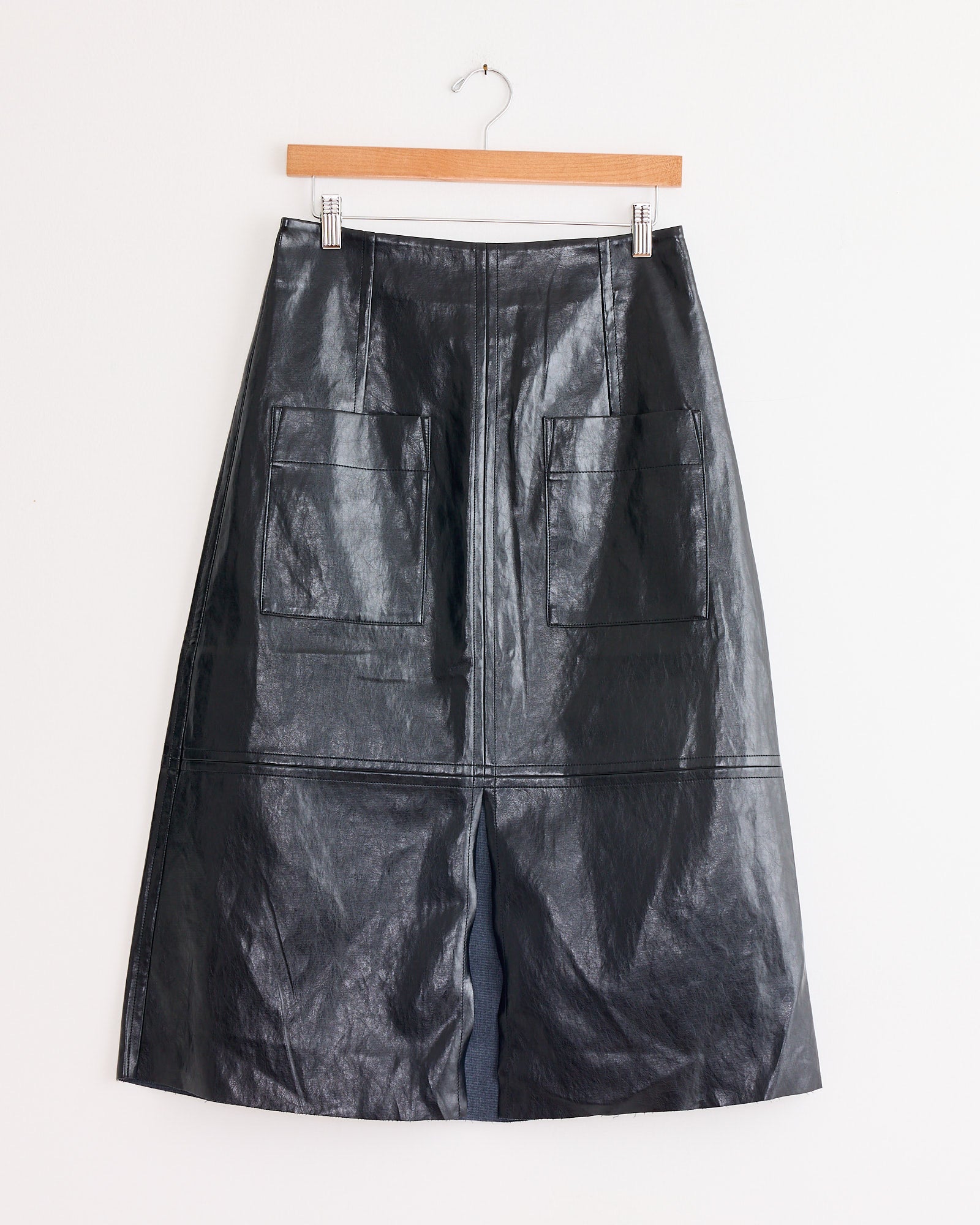 The Mijeong Park Faux Leather Skirt in Black, featuring a shiny finish and two large front pockets, is displayed on a wooden hanger against a white wall.