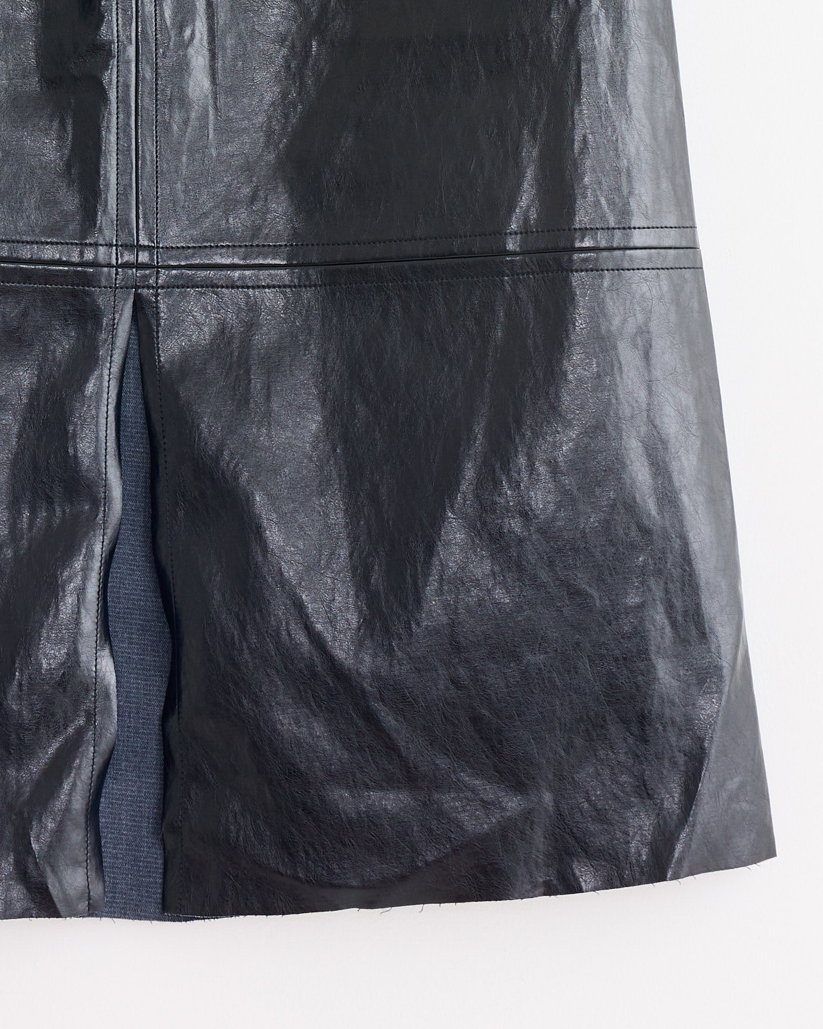 Close-up of the Mijeong Park Faux Leather Skirt in Black, featuring visible stitching and a small vertical slit on the left side. The shiny skirt is shown against a plain, light background.
