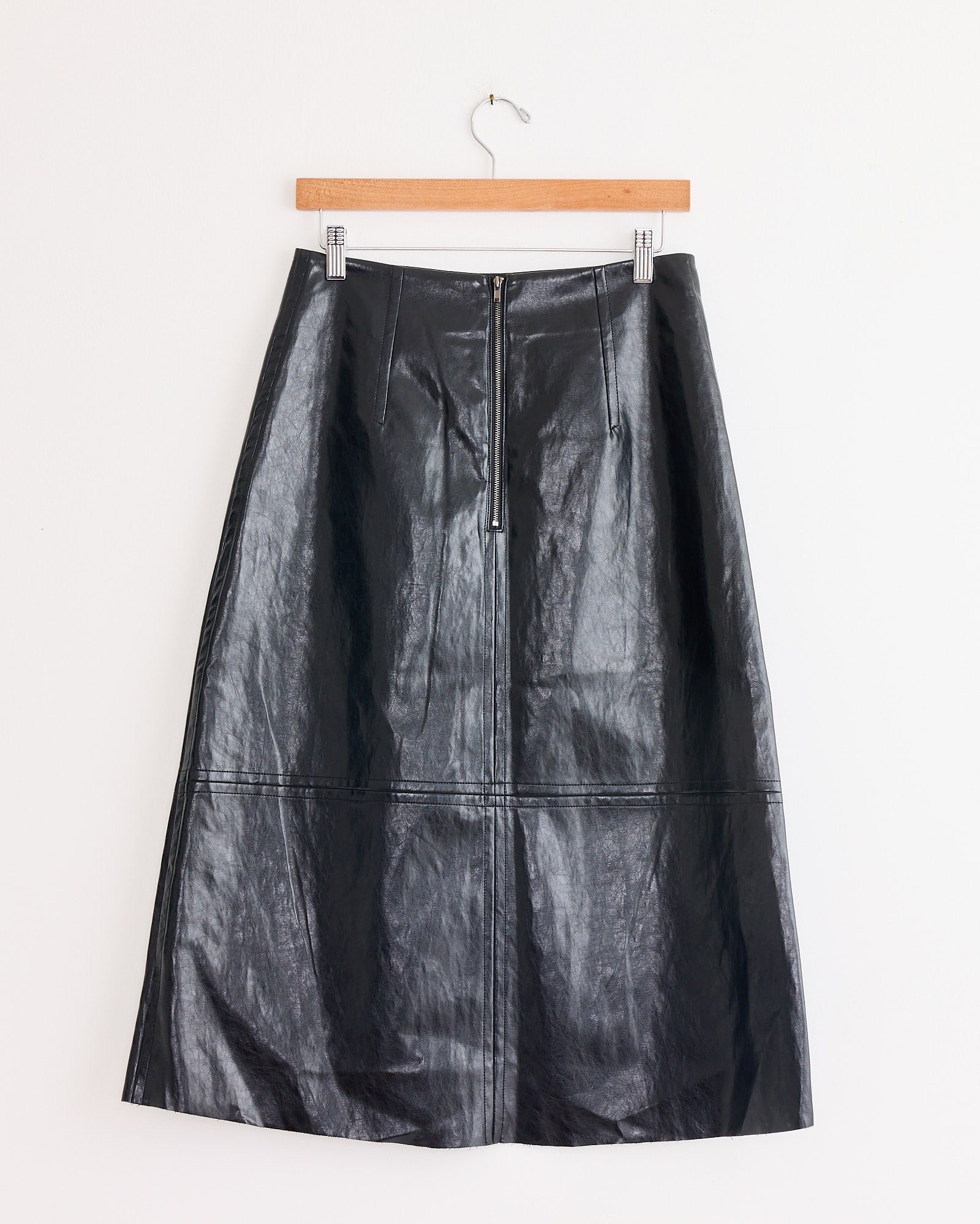 The Mijeong Park Faux Leather Skirt in Black is a long, high-waisted skirt with a front zipper, displayed on a wooden hanger against a white wall.