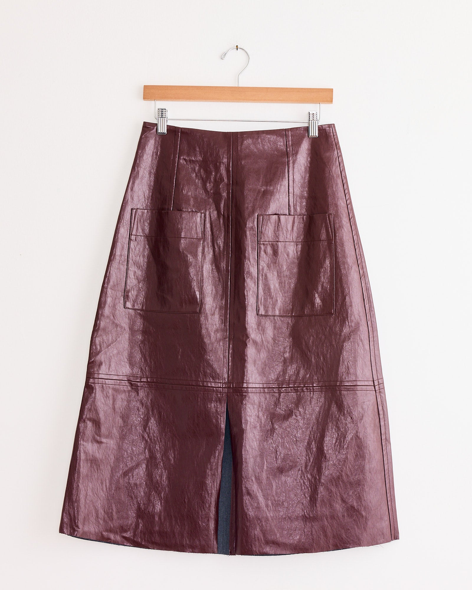 The Mijeong Park Faux Leather Skirt in Burgundy, with an A-line silhouette, features large front patch pockets and a front slit near the hem. It is displayed on a wooden hanger against a white background.