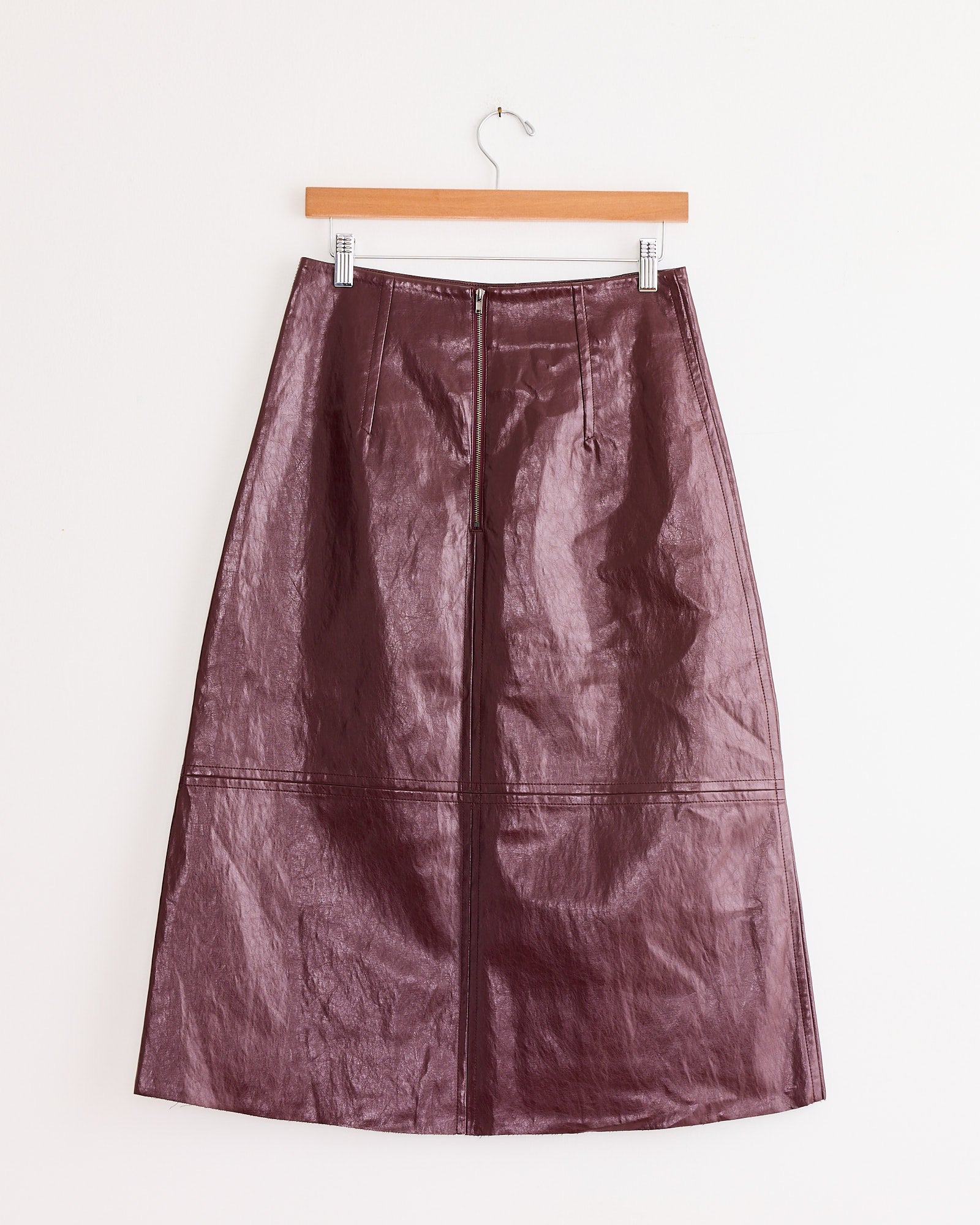 The Mijeong Park Faux Leather Skirt in Burgundy features an A-line silhouette with vertical and horizontal stitching, shown hanging on a wooden hanger against a white wall.