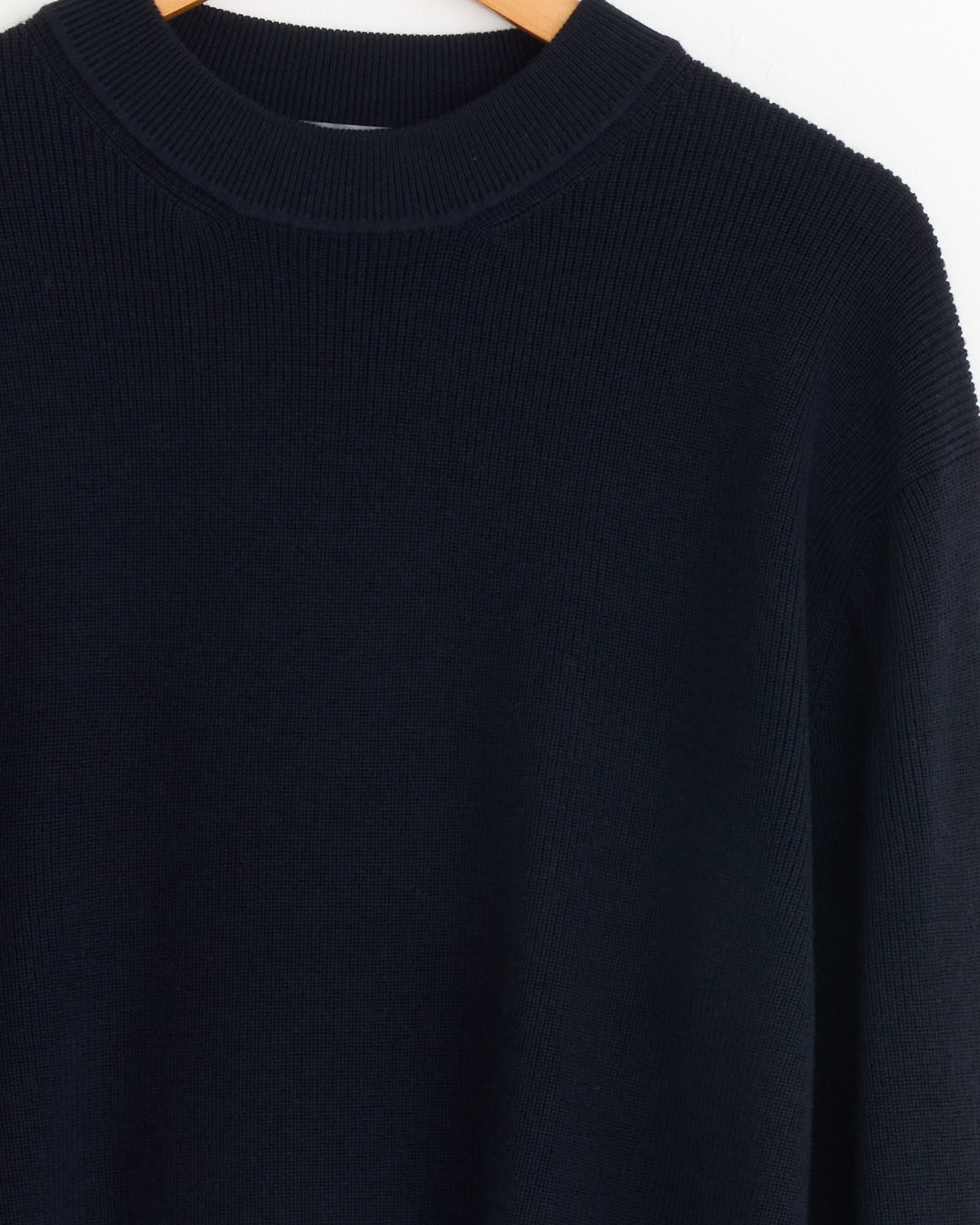 A close-up of the Margaret Howell Fine Rib Merino Wool Crewneck in Ink, featuring a relaxed fit, hangs on a wooden hanger against a white background.