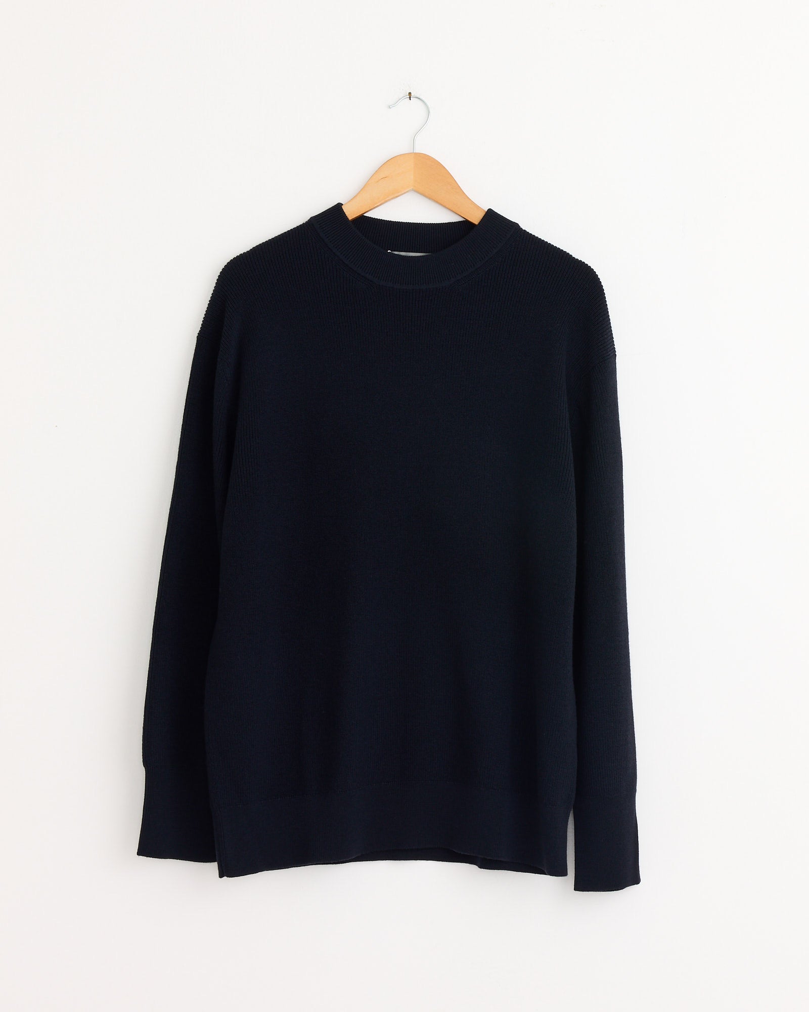 The Fine Rib Merino Wool Crewneck in Ink by Margaret Howell, featuring a relaxed fit and long sleeves, is shown on a wooden hanger against a white wall.