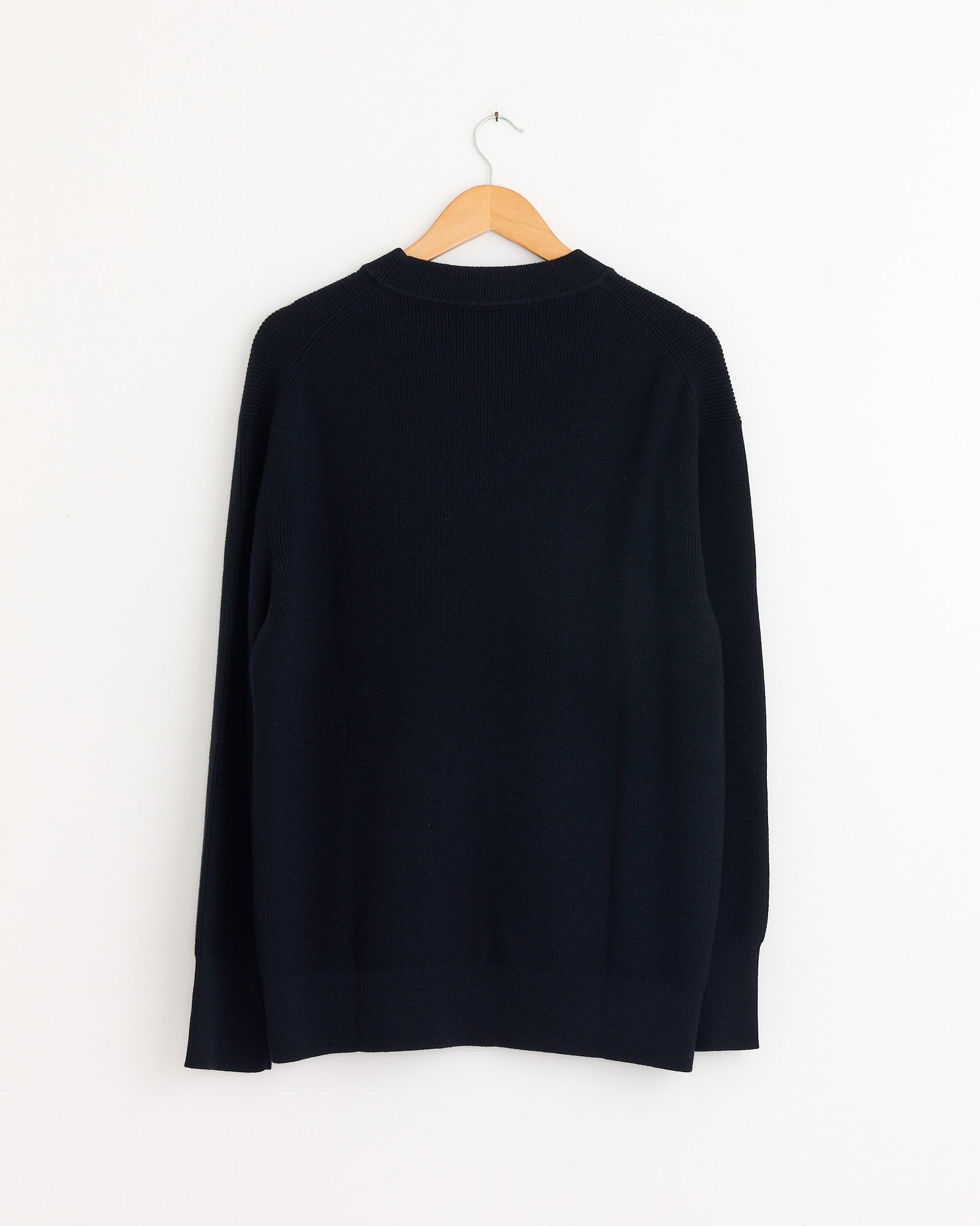 The Margaret Howell Fine Rib Merino Wool Crewneck in Ink, shown from the back, features a relaxed fit and hangs on a wooden hanger against a white wall.