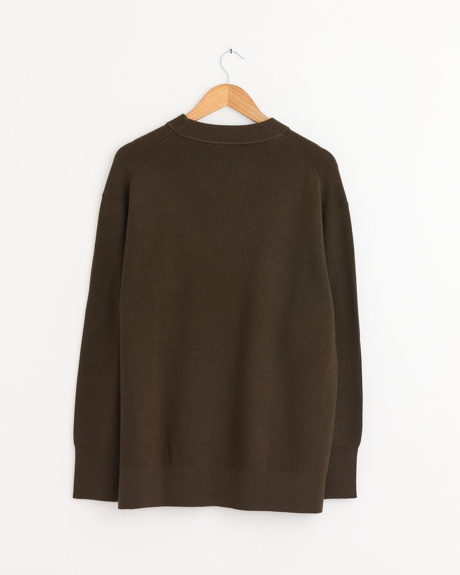 The Margaret Howell Fine Rib Merino Wool Crewneck in Khaki is shown from the back on a wooden hanger, against a plain white wall. It has a relaxed fit with ribbed collar, cuffs, and hem.