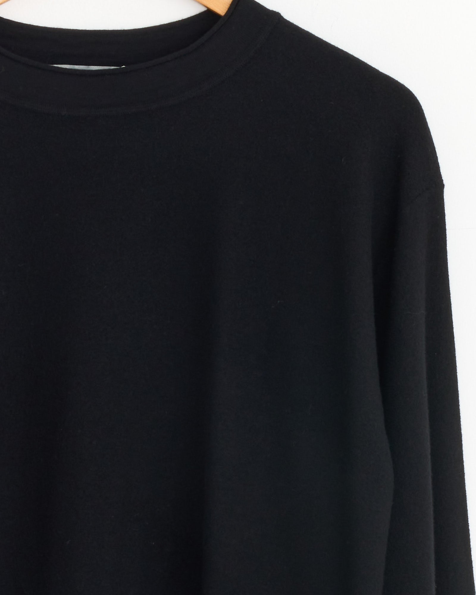 The Margaret Howell Felted Merino Crewneck in Black, made from lightweight felted wool, features a classic crew neck and long sleeves. It is displayed on a wooden hanger against a white background.