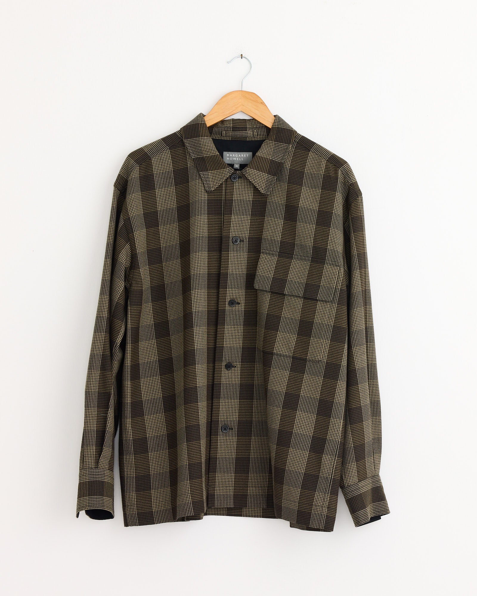 The Margaret Howell Flap Pocket Overshirt in Black/Brown/Multi, a long-sleeved, collared button-up in plaid cotton wool twill, hangs on a wooden hanger against a plain white wall.