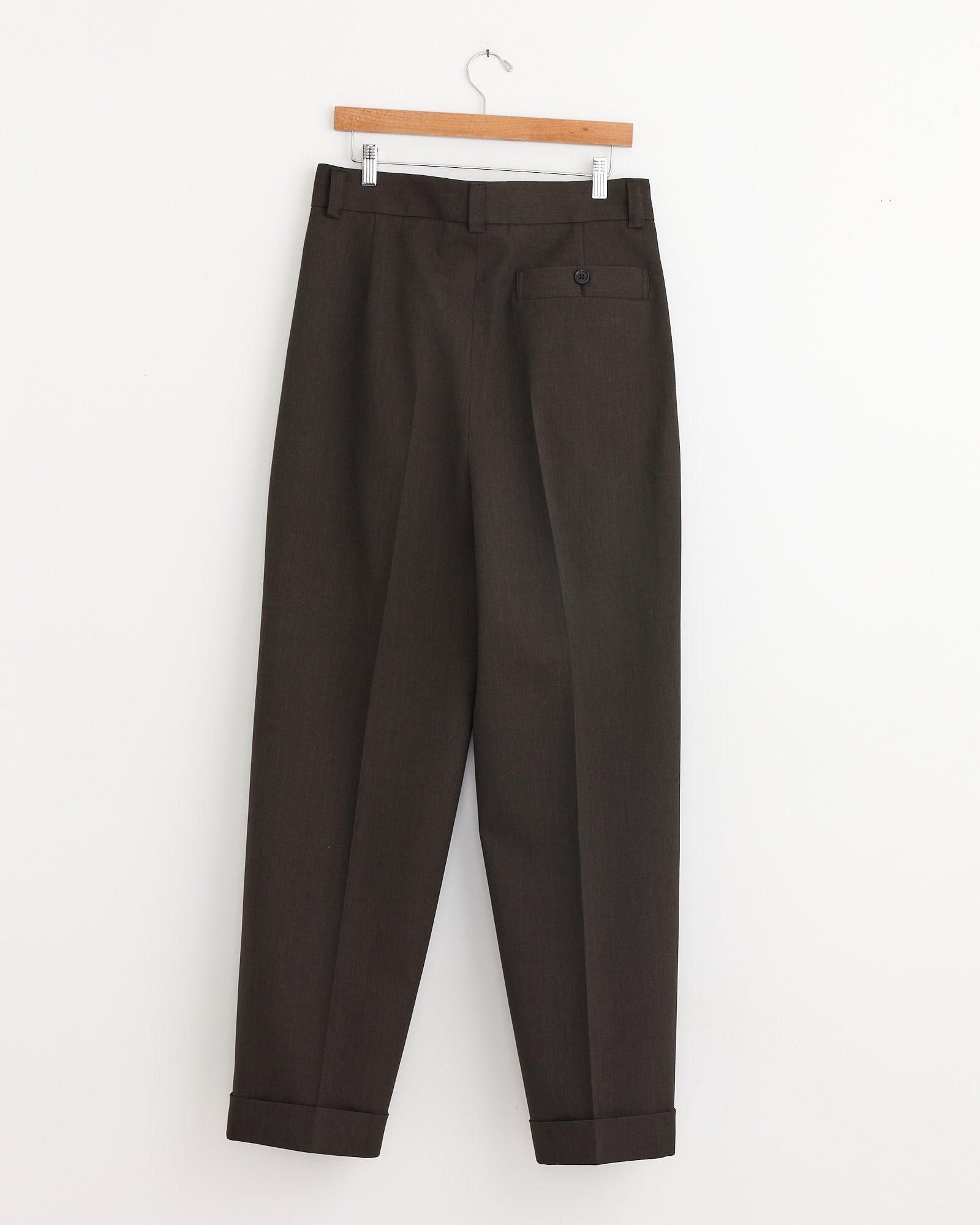 The Stitch Pleat Trouser in Khaki Brown by Margaret Howell, featuring a cuffed hem, hangs on a wooden hanger against a plain white wall.
