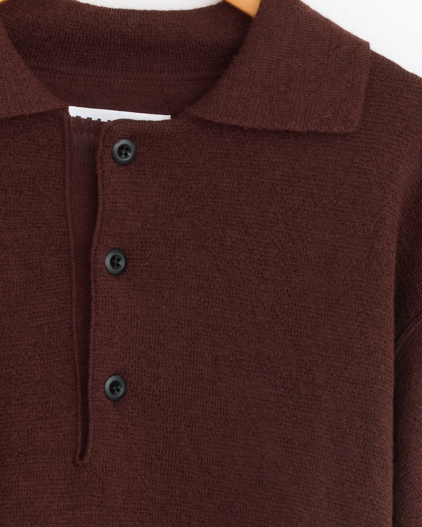 Close-up of the MHL by Margaret Howell Oversized Knitted Polo in Dark Brown, featuring a collar and three black buttons, displayed on a hanger against a white background. For fit or sizing questions, contact us via online chat.