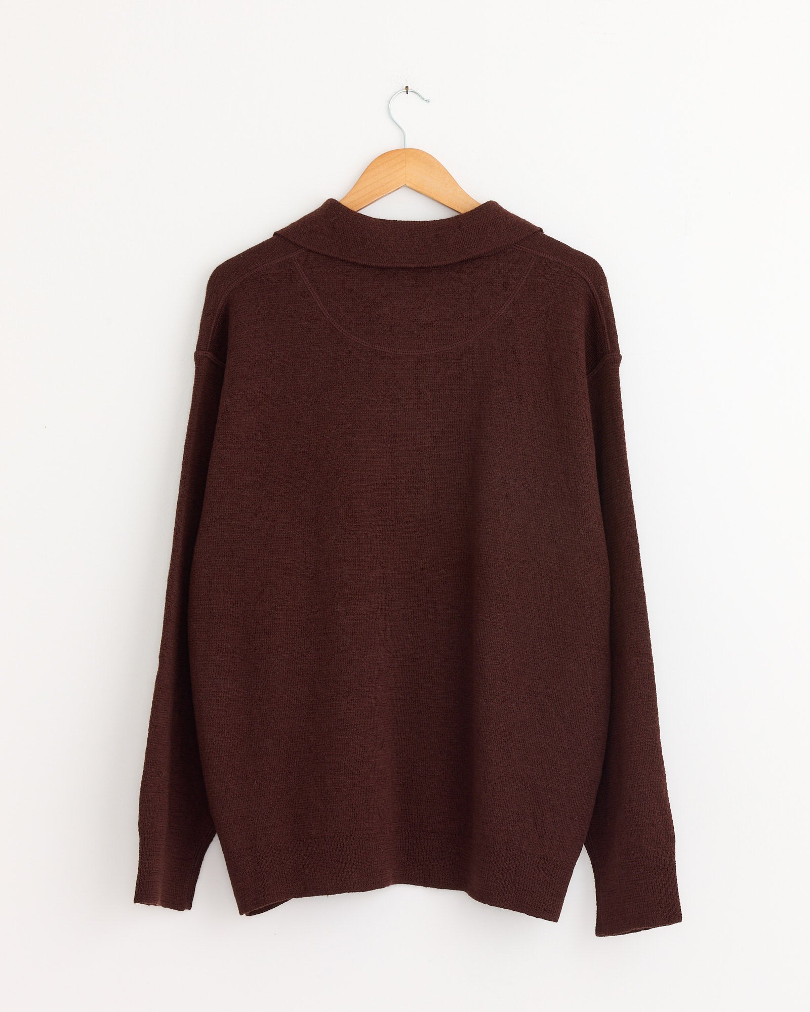 The Oversized Knitted Polo in Dark Brown by MHL by Margaret Howell hangs from a wooden hanger against a plain white wall, shown from the back—perfect for lovers of classic style and easy sizing. Chat online for fit questions.